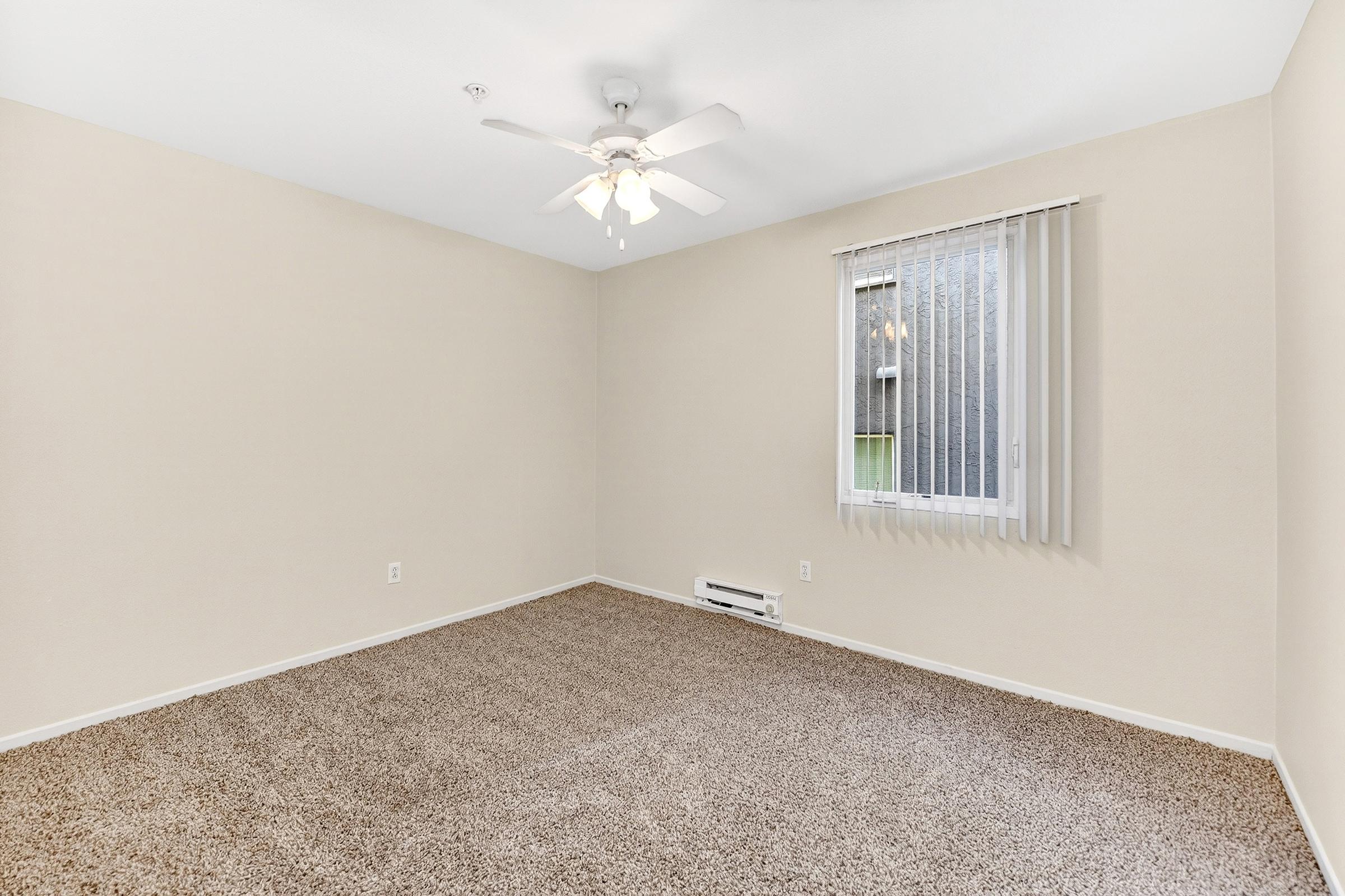 A spacious, empty room with light beige walls and a ceiling fan. The floor is covered in beige carpet, and there's a window with vertical blinds allowing natural light. A heating unit is near the baseboard. The space appears clean and ready for furnishing.