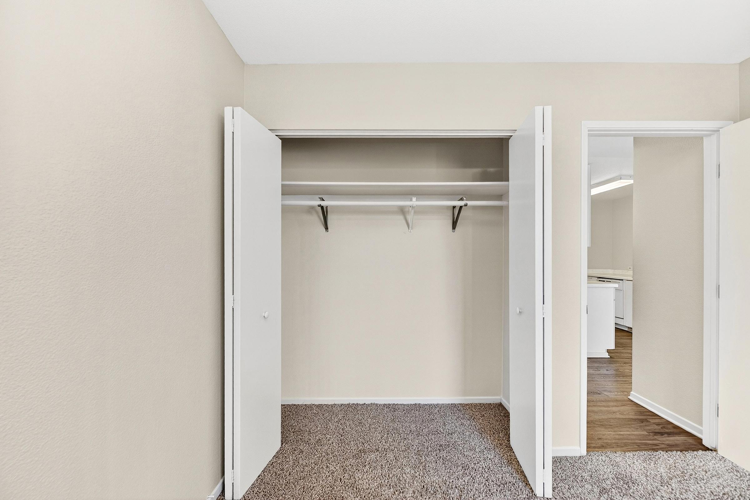 A clean, empty closet with open white double doors, featuring a single shelf and hanging rods. The walls are painted a soft beige, and the floor is covered with beige carpet. In the background, a glimpse of a bright kitchen can be seen through an adjoining open doorway.