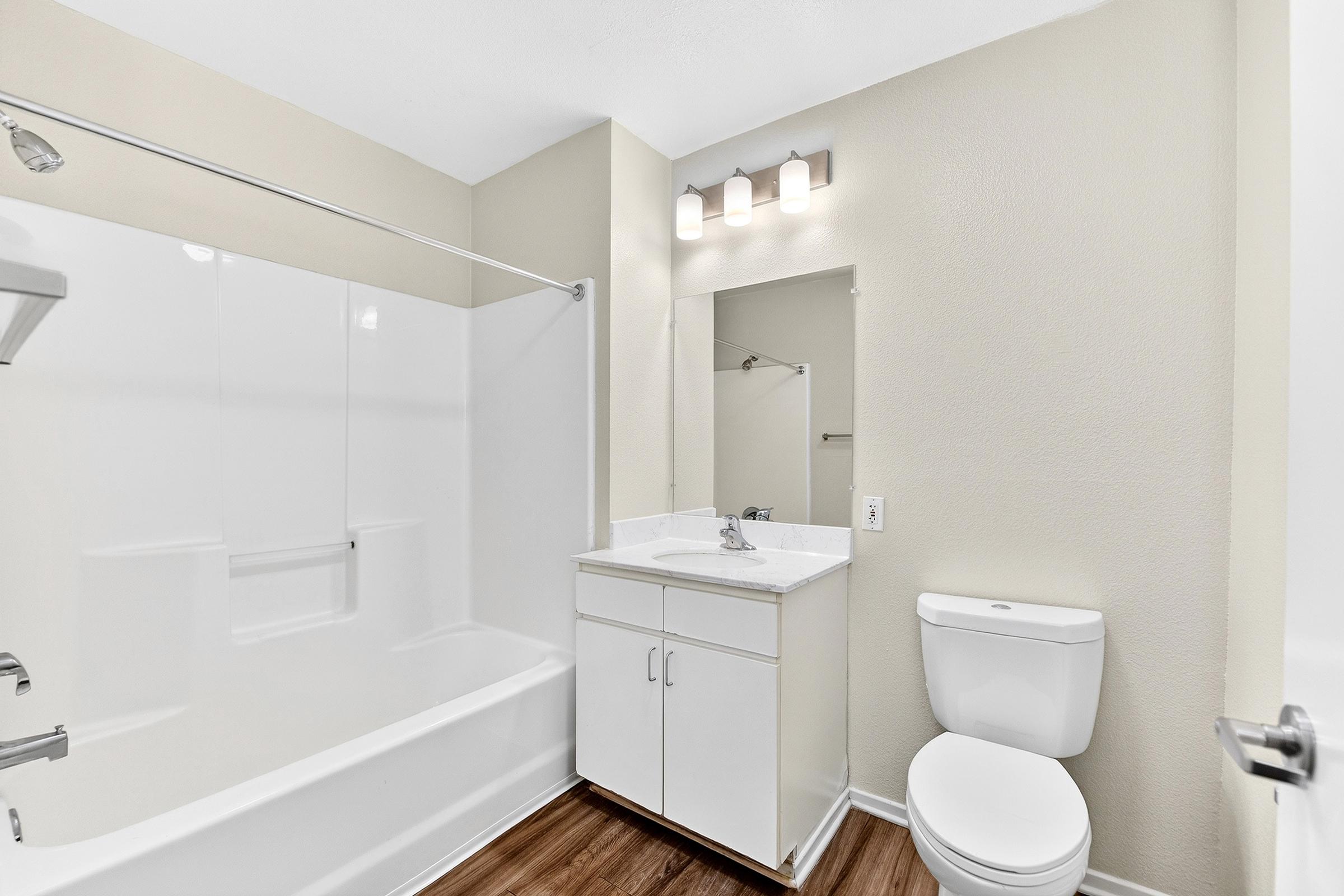 A clean, modern bathroom featuring a white bathtub with a glass shower door, a sink with a white cabinet beneath, a toilet, a large mirror, and overhead lighting. The walls are painted a light beige, and the floor is covered with natural wood-style laminate.