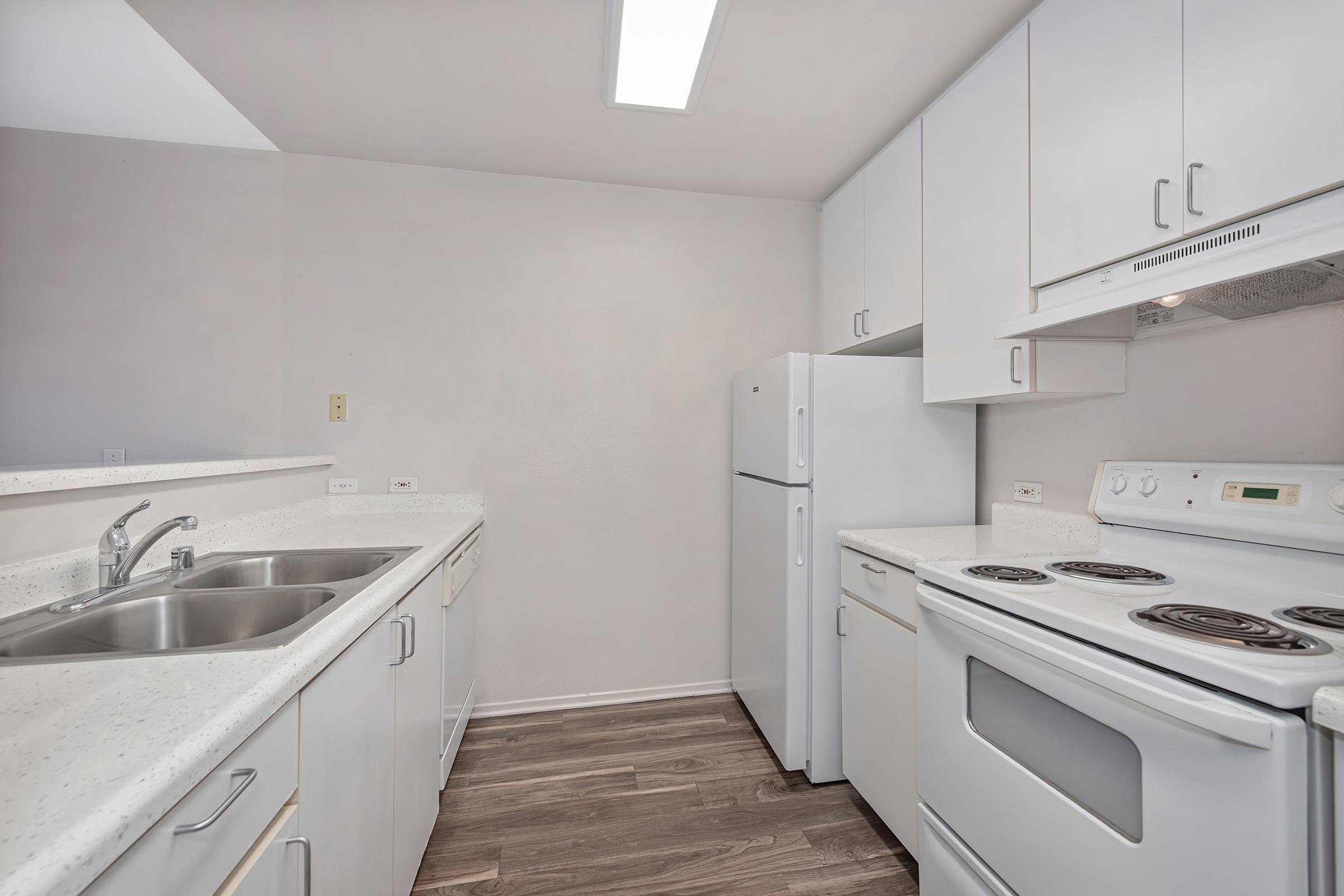 A modern kitchen featuring white cabinetry, a double sink, a refrigerator, and a stove with an oven. The countertops are light-colored with a smooth finish, and the flooring is wood-like. Natural light comes from a ceiling fixture, creating a bright and inviting space.