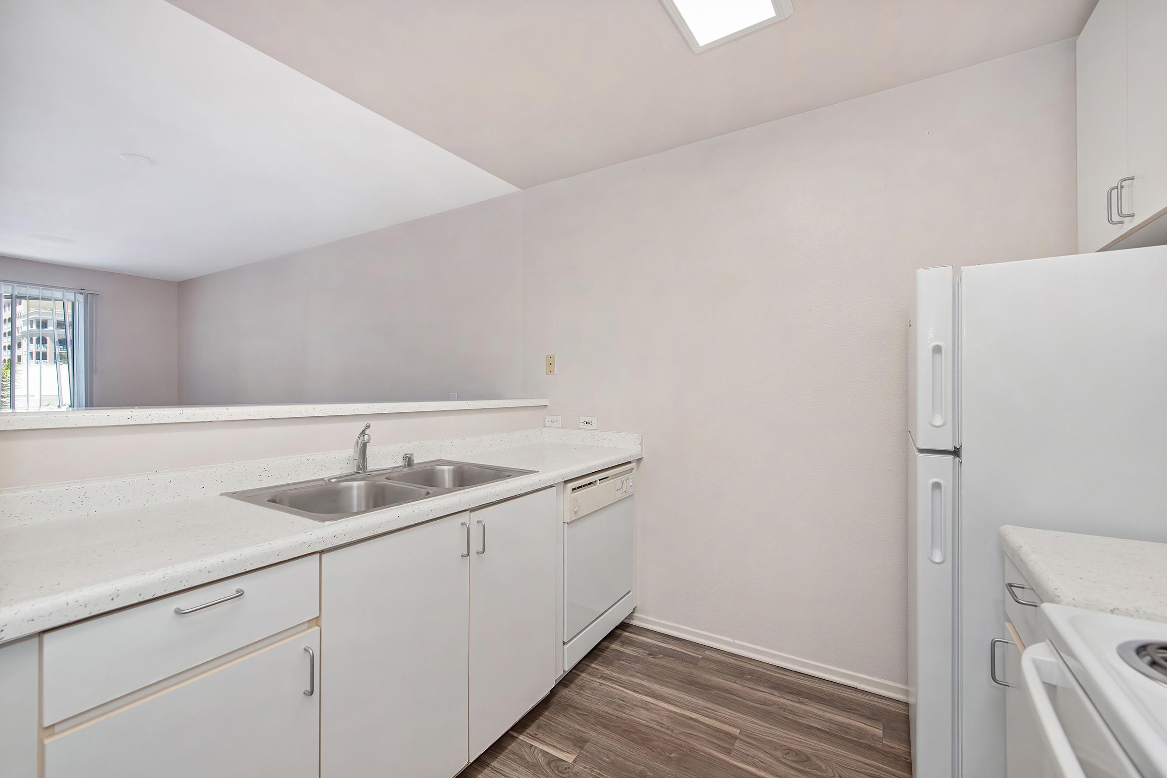 A modern kitchen featuring a double sink, dishwasher, and a white refrigerator. The cabinetry is light-colored, and the countertops are a clean, speckled white. The floor is wood-like laminate. Natural light enters from a window, creating a bright and inviting atmosphere.