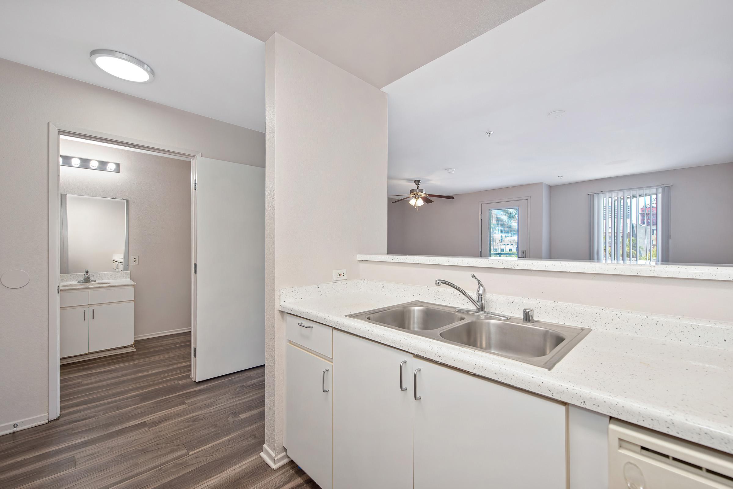 Interior view of a modern kitchen featuring a double sink, light countertops, and dark wood flooring. A doorway leads to a bathroom area with a mirror, and a ceiling fan is visible in the background. Natural light comes in through a window, creating a bright and open atmosphere.