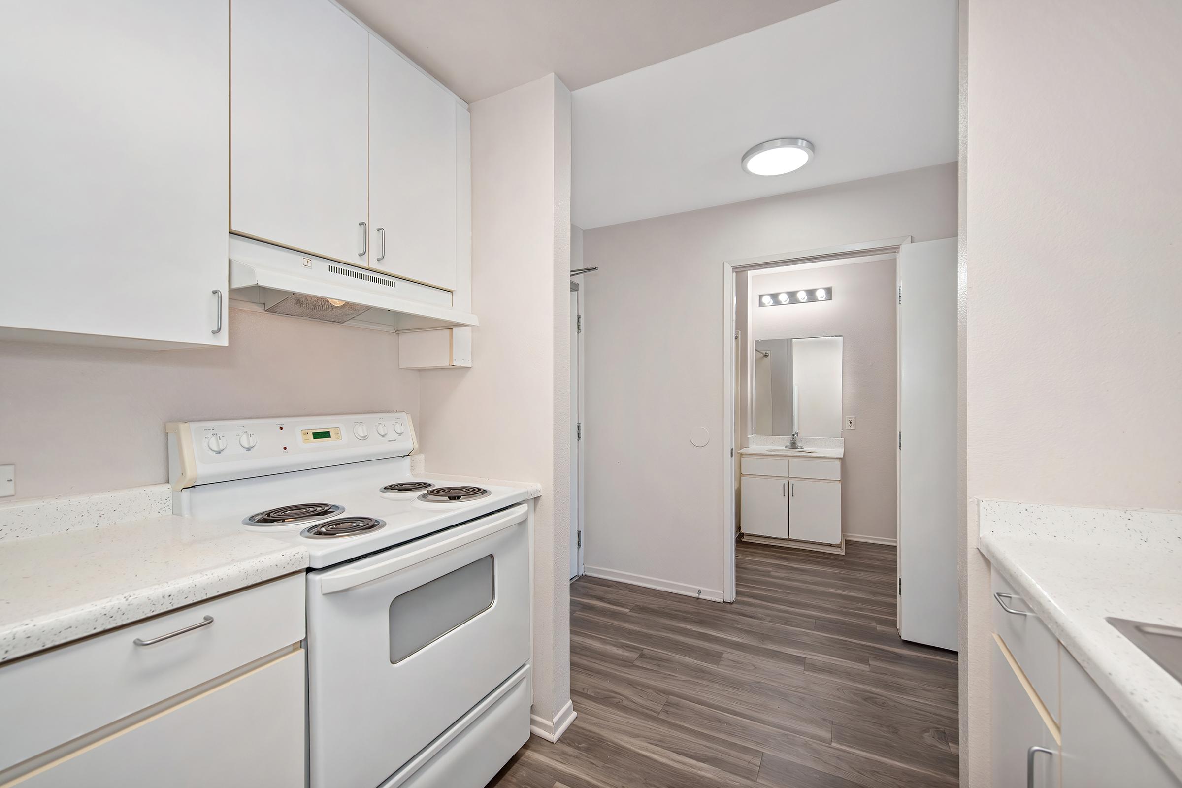A modern kitchen with white cabinetry, a white stove and oven, and a light countertop. Wood laminate flooring runs throughout the space. A doorway leads to a bathroom area with bright lighting, adding to the open and airy feel of the kitchen.