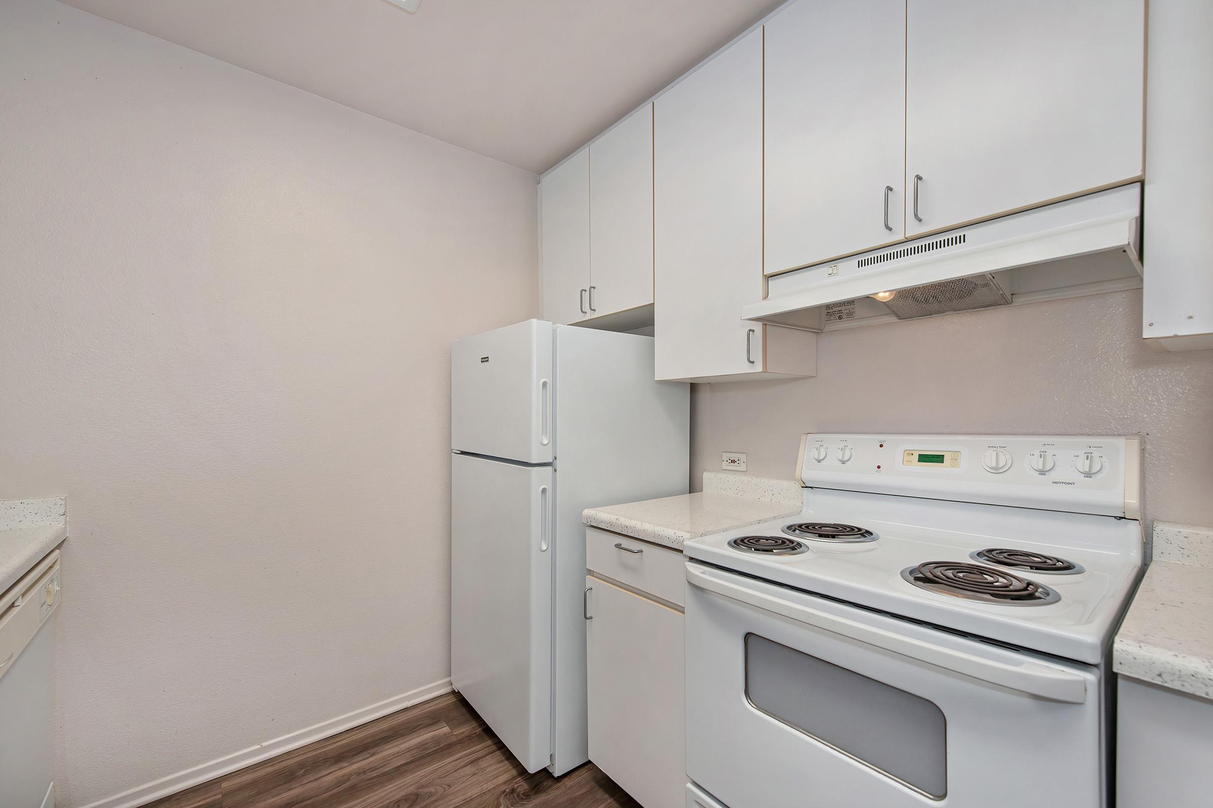 A bright and clean kitchen featuring white cabinetry, a white refrigerator, and a white stove with an oven. The countertops are light-colored, and the flooring is a warm wood tone, creating a modern and inviting atmosphere. Walls are painted in a soft, neutral hue.