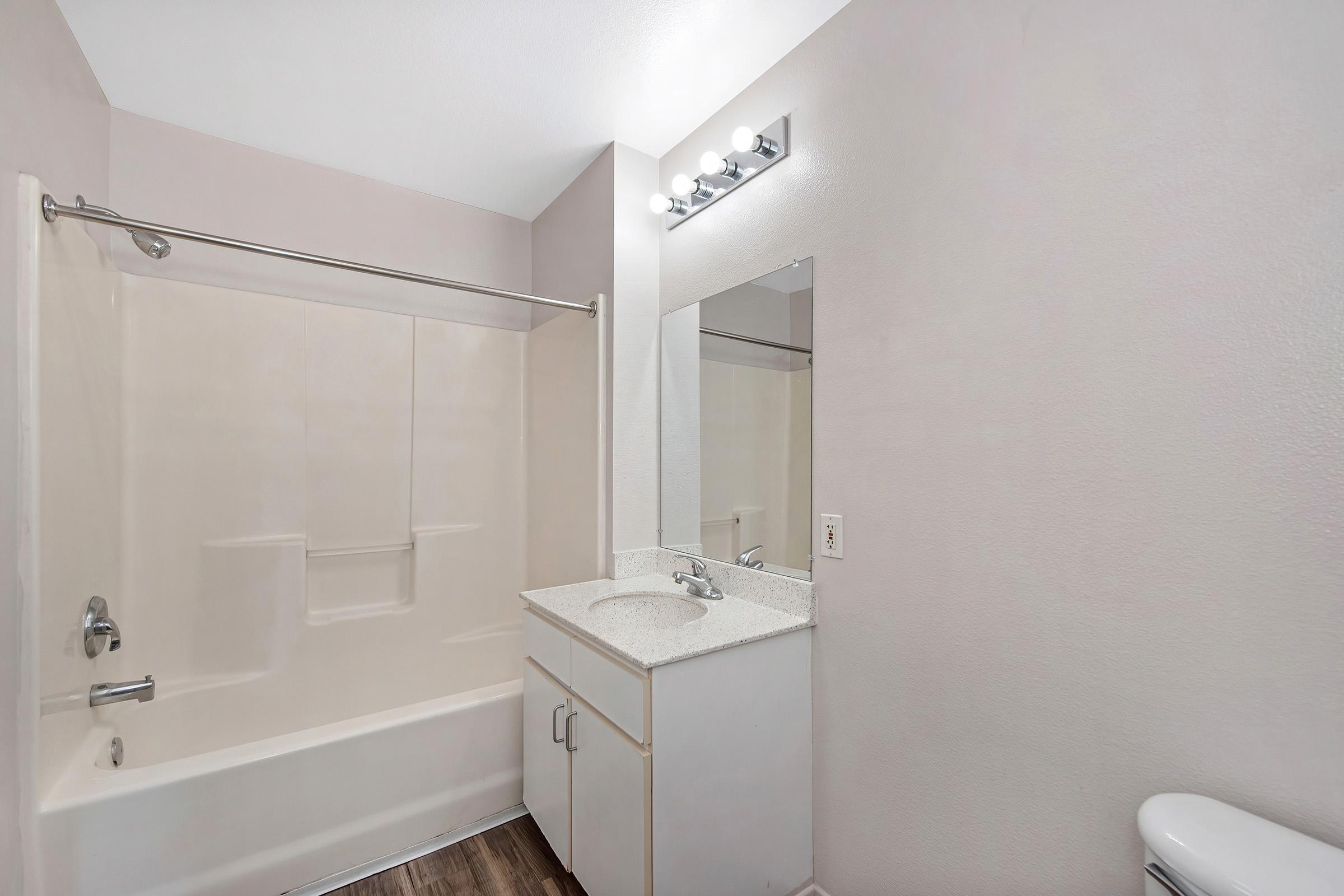 A clean and modern bathroom featuring a bathtub with a shower, a small vanity with a sink, a large mirror above it, and light fixtures. The walls are painted in a soft, neutral color, and the flooring is a wood-like laminate, creating a bright and inviting space.