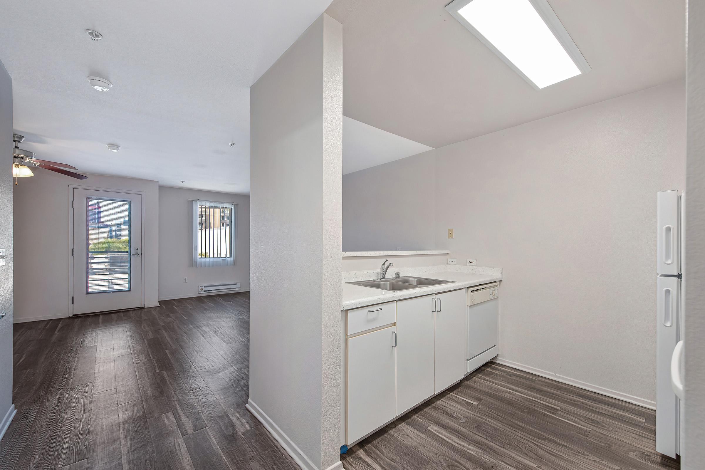 Interior view of a modern apartment featuring an open layout with a kitchen on the right, equipped with a sink, dishwasher, and refrigerator. The living area has large windows with natural light and sliding patio doors, along with sleek wooden flooring and neutral-colored walls.