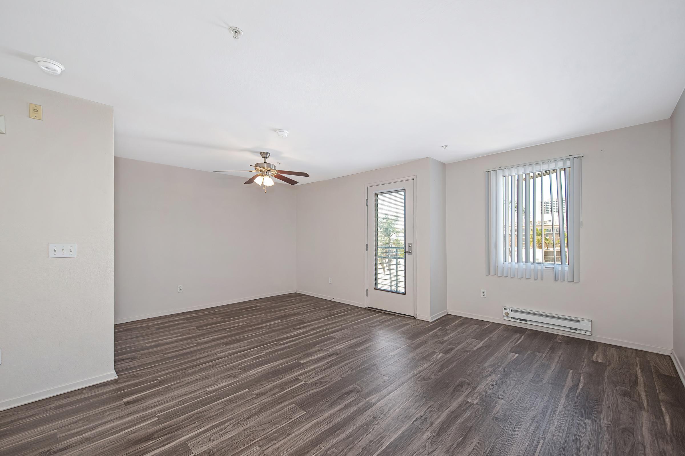 A spacious, empty room with light gray walls and a wood-like floor. A ceiling fan is installed, and there is a sliding glass door leading to a small balcony. Vertical blinds on the window allow natural light to enter, creating a bright and airy atmosphere.