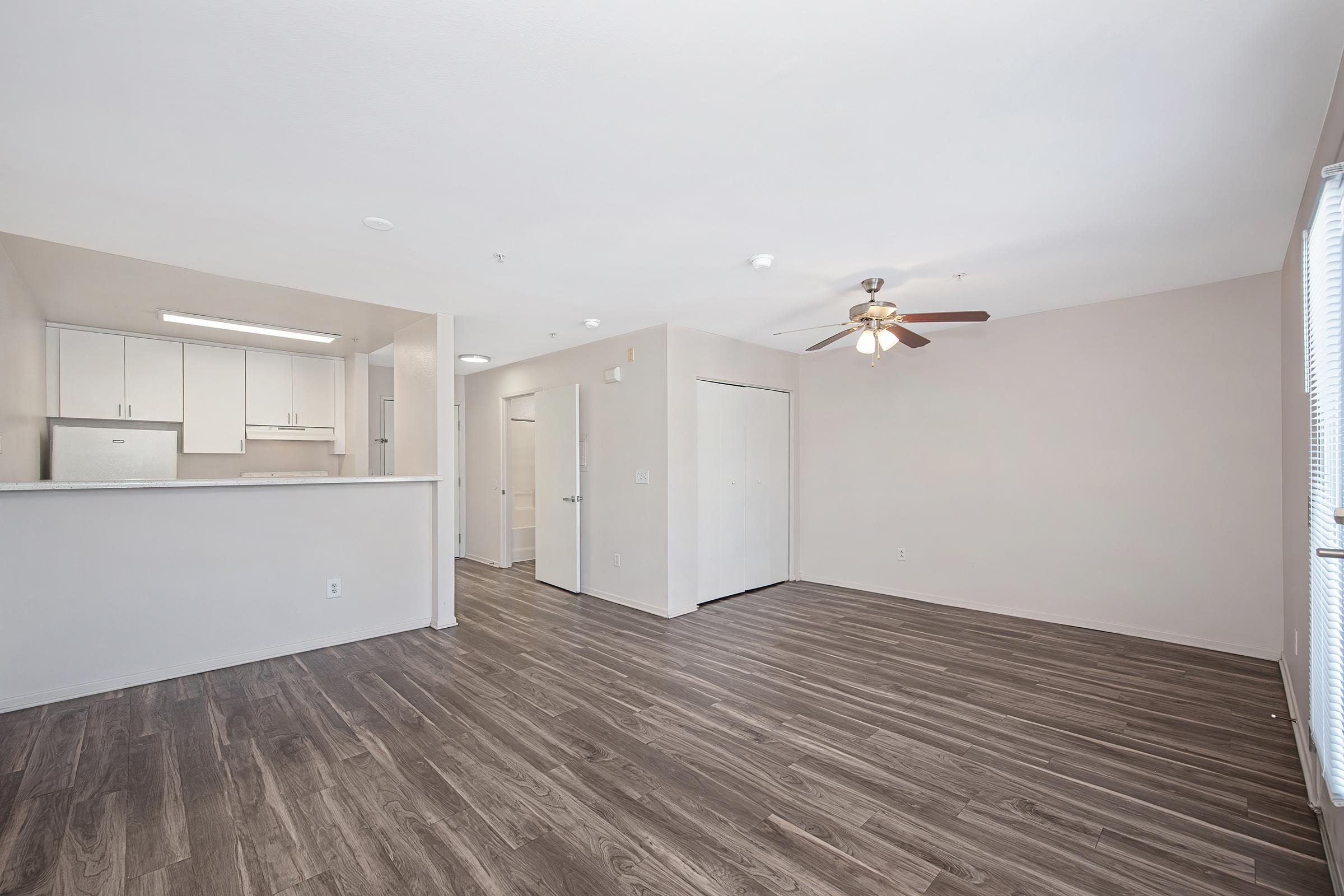 A spacious interior of an apartment featuring light-colored walls, wooden flooring, a ceiling fan, and an open kitchen area. The layout includes a doorway leading to another room, with natural light streaming in through a window. The overall atmosphere is bright and inviting.