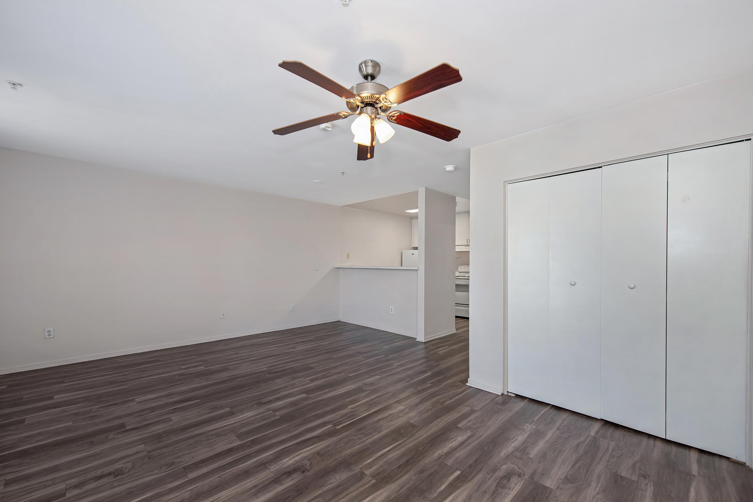 A spacious living area featuring light-colored walls, a ceiling fan with wooden blades, and contemporary flooring. To the right, there are two closed white closet doors, and in the background, there's an open kitchen area with visible appliances. Natural light brightens the space, creating a welcoming atmosphere.