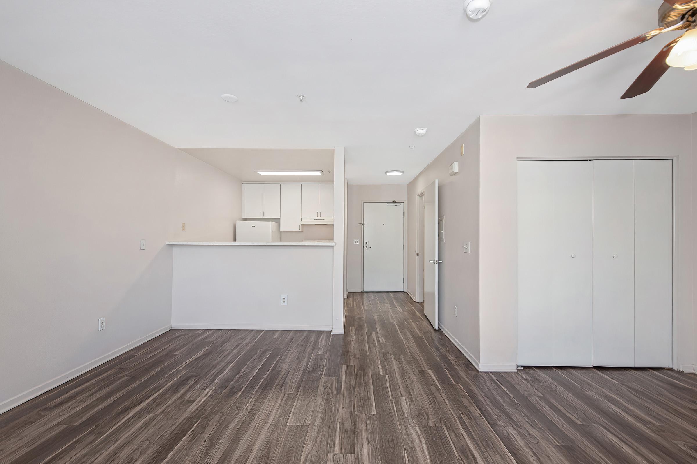 Spacious interior view of an apartment featuring a living area with wooden flooring, a ceiling fan, and an open kitchen in the background. The kitchen has white cabinets, and there are doors leading to other rooms. Natural light fills the space, creating a bright and welcoming atmosphere.