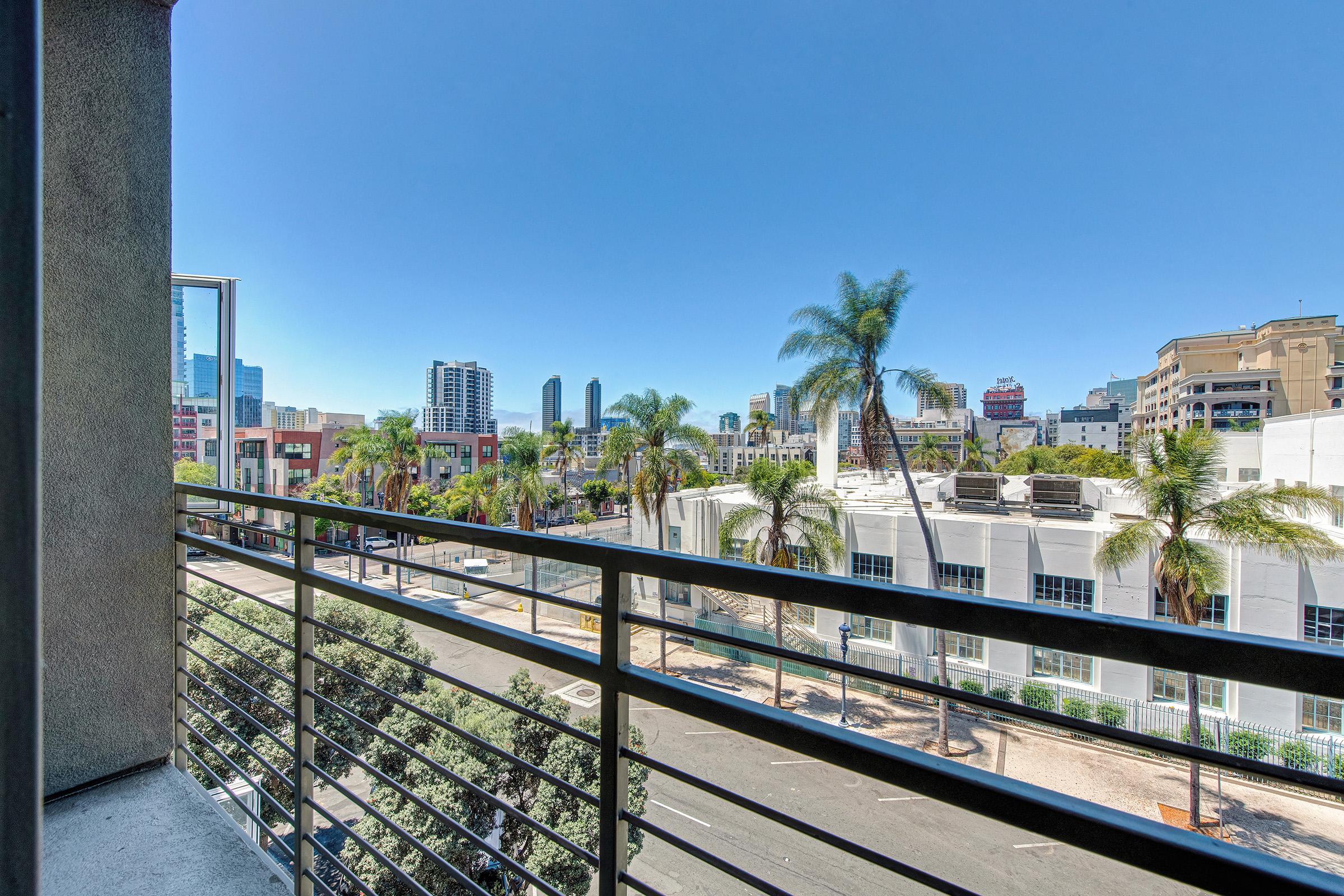 View from a balcony overlooking a city street lined with palm trees, with tall buildings in the background. The sky is clear and bright blue, indicating a sunny day. The scene captures a vibrant urban landscape.