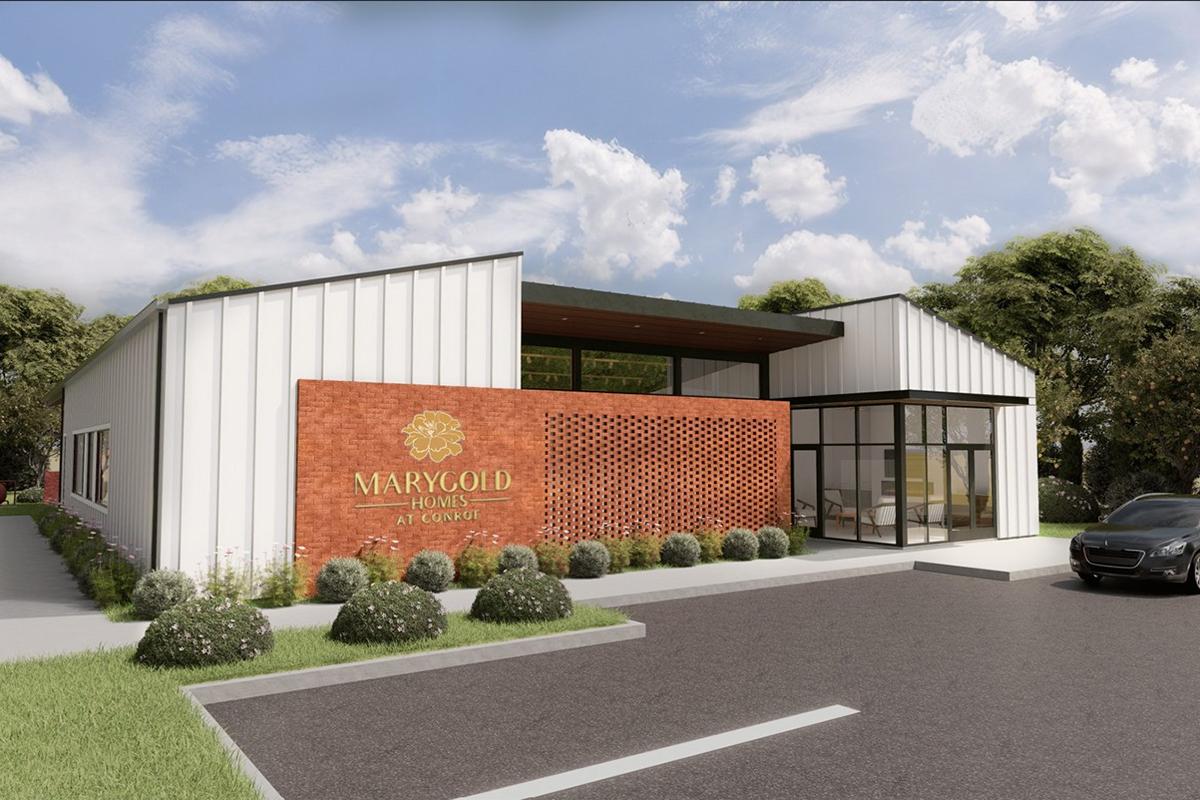 Modern building exterior of Marygold Homes, featuring a mix of brick and metal materials. The design showcases large windows and a welcoming entrance. Surrounding landscaping includes shrubs and trees, with a paved parking area visible in the foreground under a blue sky with fluffy clouds.
