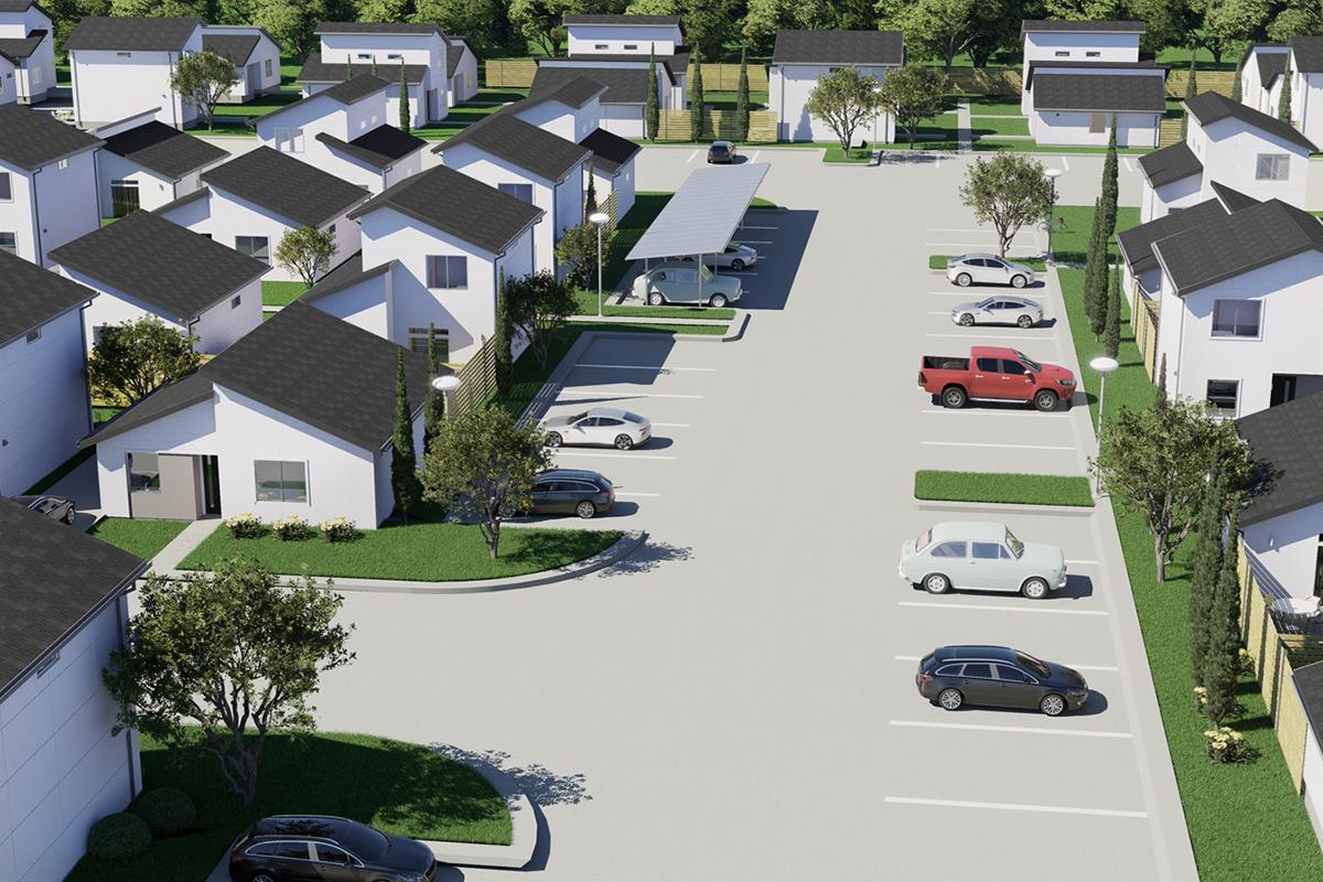 Aerial view of a residential neighborhood featuring modern houses with pitched roofs. A spacious parking lot is visible, with several parked cars and a covered parking area. Surrounding greenery includes trees and shrubbery, adding a touch of nature to the community layout.