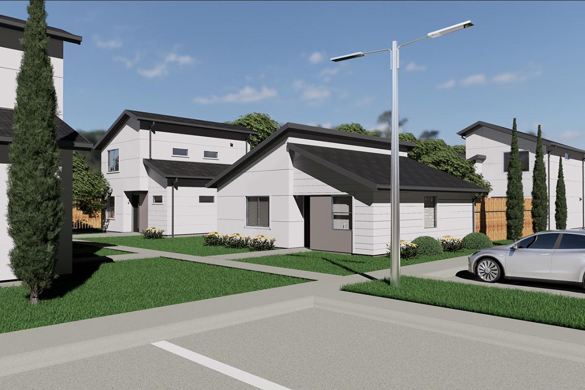 A modern residential community featuring several two-story and single-story houses with flat roofs. The buildings are surrounded by greenery, including neatly trimmed lawns and flowering plants. A streetlight stands along the walkway, and a parked car is visible in the foreground. The sky is clear and blue.
