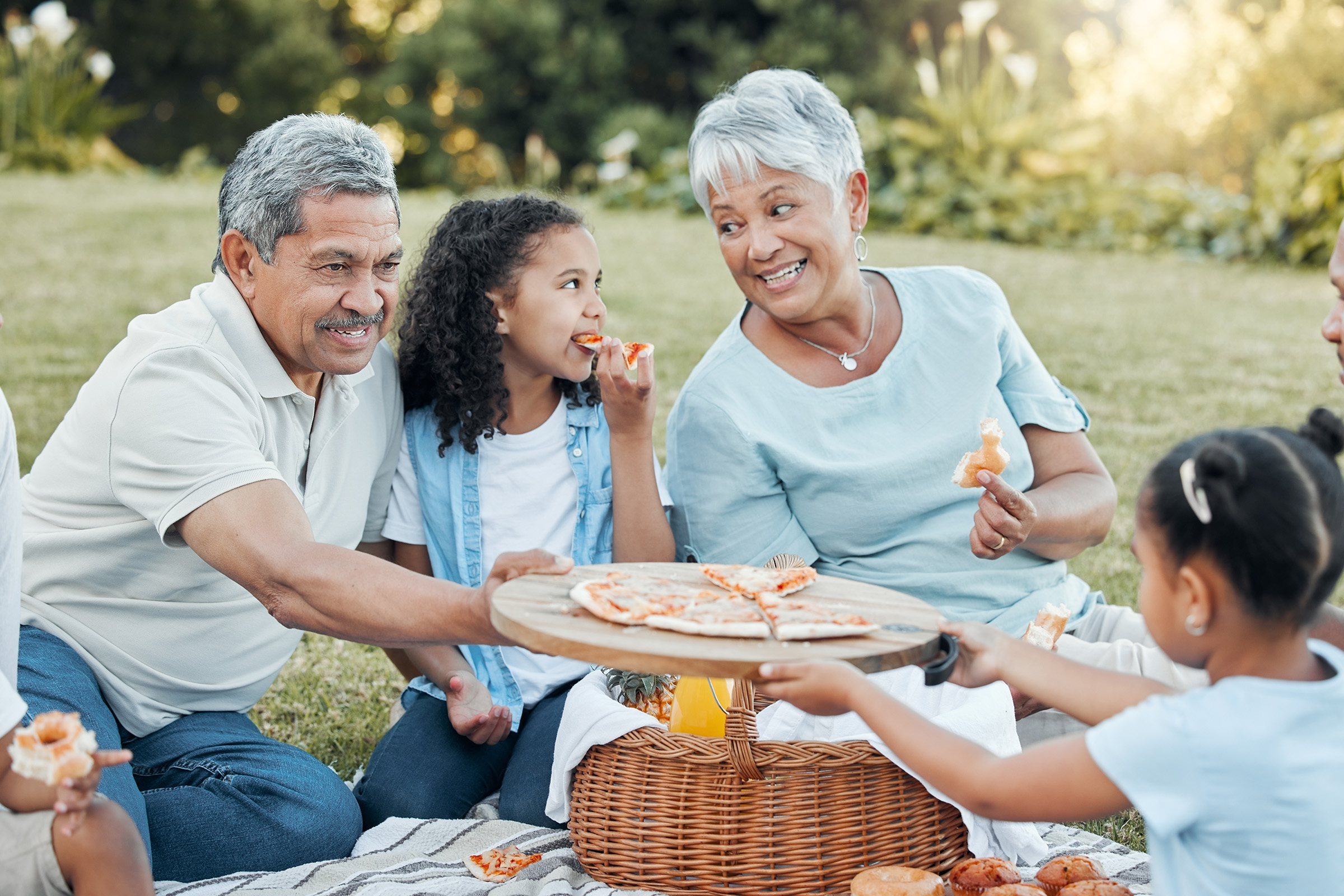 A joyful family picnic scene in a park. An older couple shares snacks with their grandchildren. The grandmother smiles as she hands a pizza slice to a young girl who is enjoying her food, while the grandfather looks on affectionately. The atmosphere is relaxed and filled with laughter, surrounded by greenery.