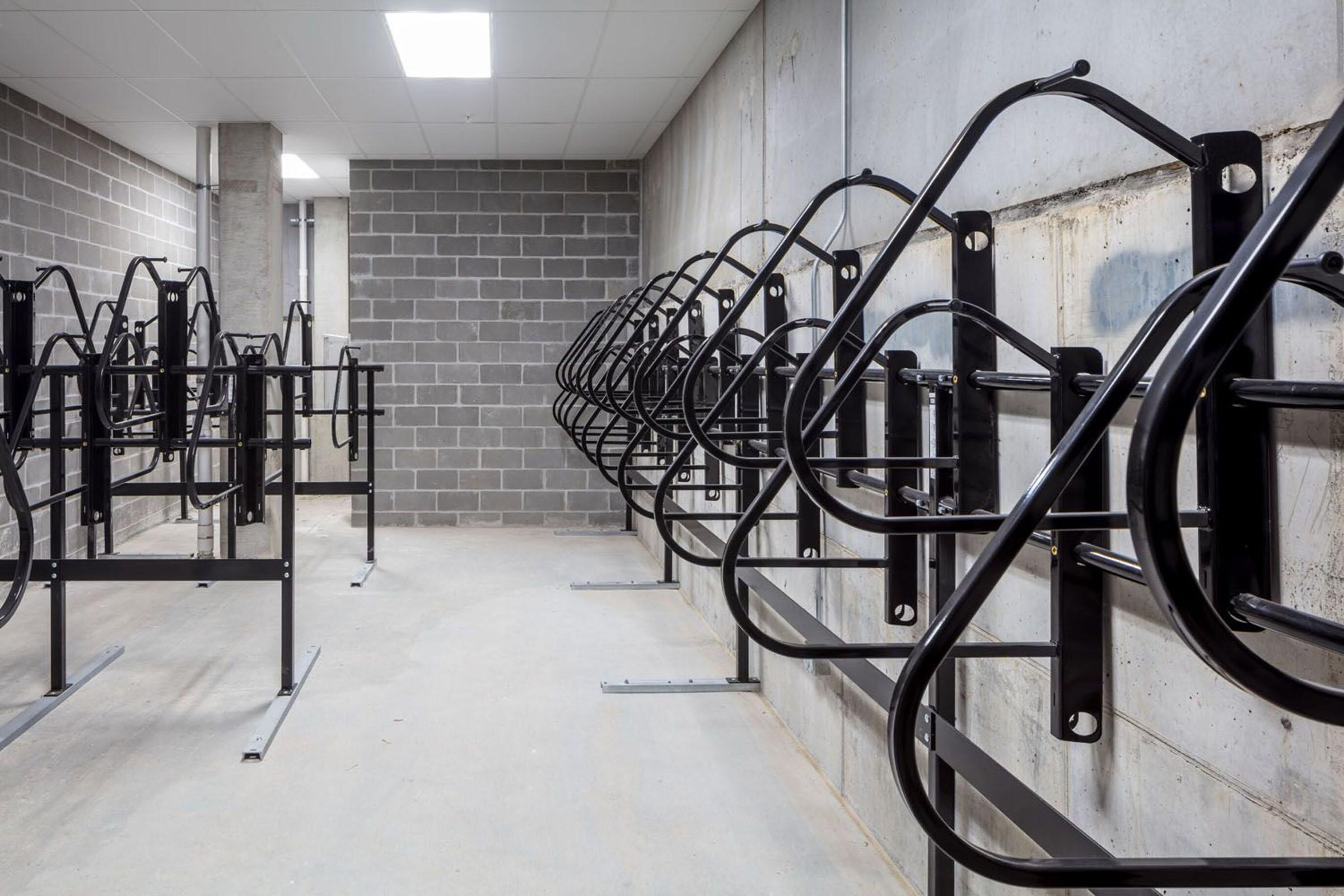A row of black metal bike racks mounted against a concrete wall in a well-lit indoor space. The racks are designed for securing bicycles, with multiple spaces created for parking them. The surrounding area features a gray concrete floor and walls, giving it a modern industrial look.