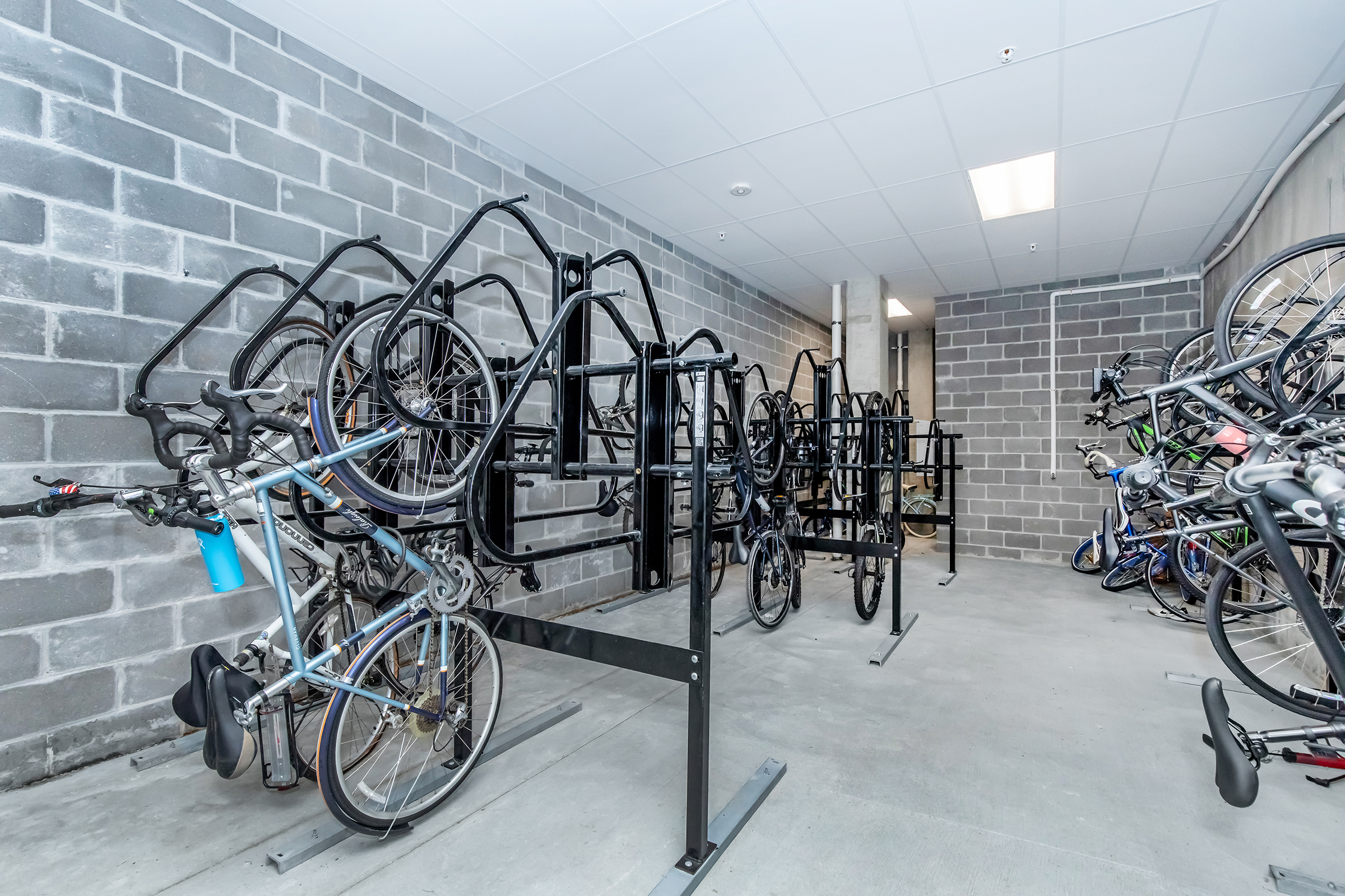Indoor bicycle storage area featuring multiple bike racks. The racks are empty and a few bicycles are secured, with a gray concrete block wall in the background. The space is well-lit with overhead lighting.