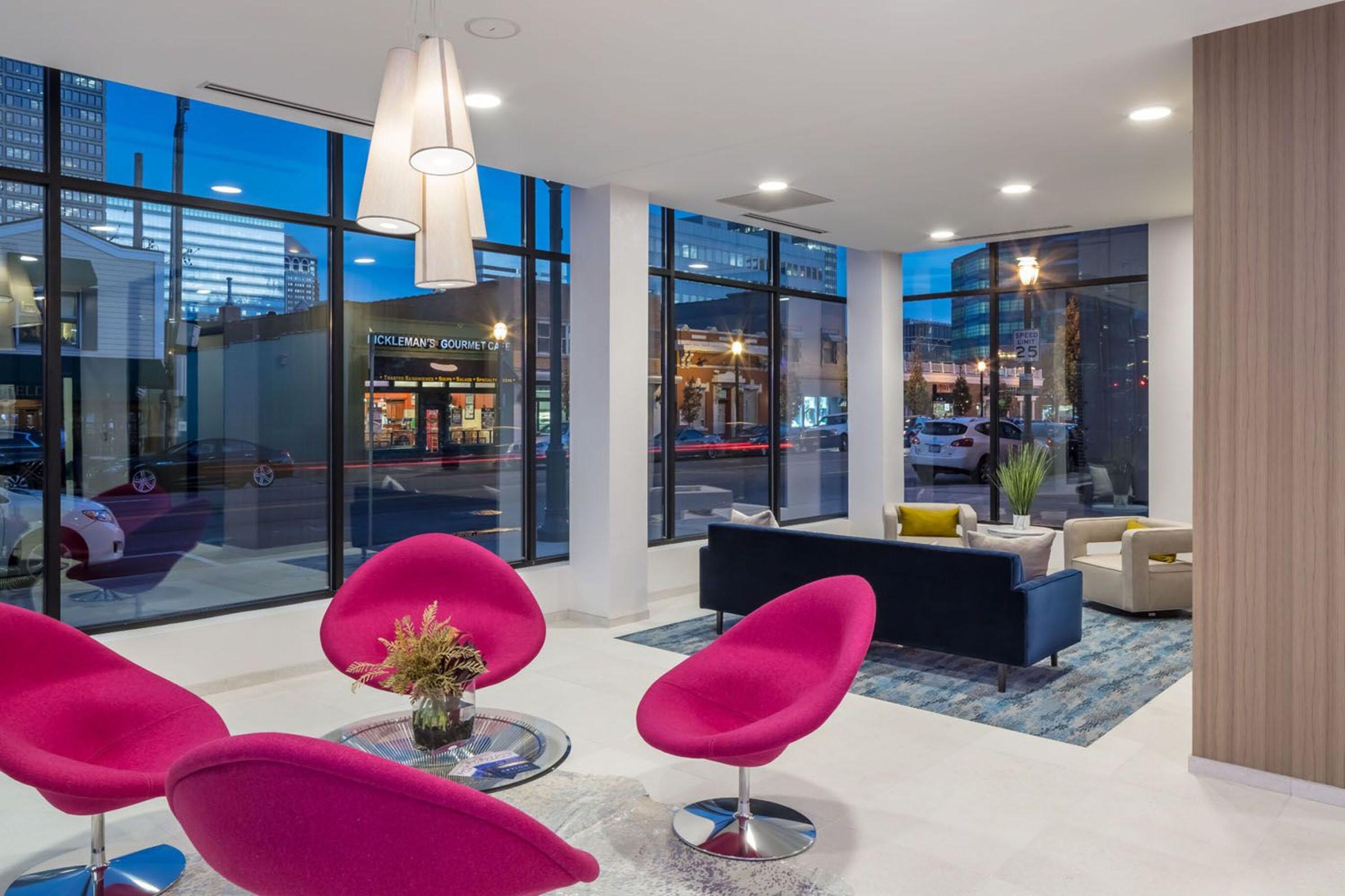 Modern interior of a lounge area featuring large windows, pink chairs, a stylish coffee table, and a dark sofa. The decor includes plants and a light fixture, with a view of a city street outside. The space combines contemporary design elements with a welcoming atmosphere.
