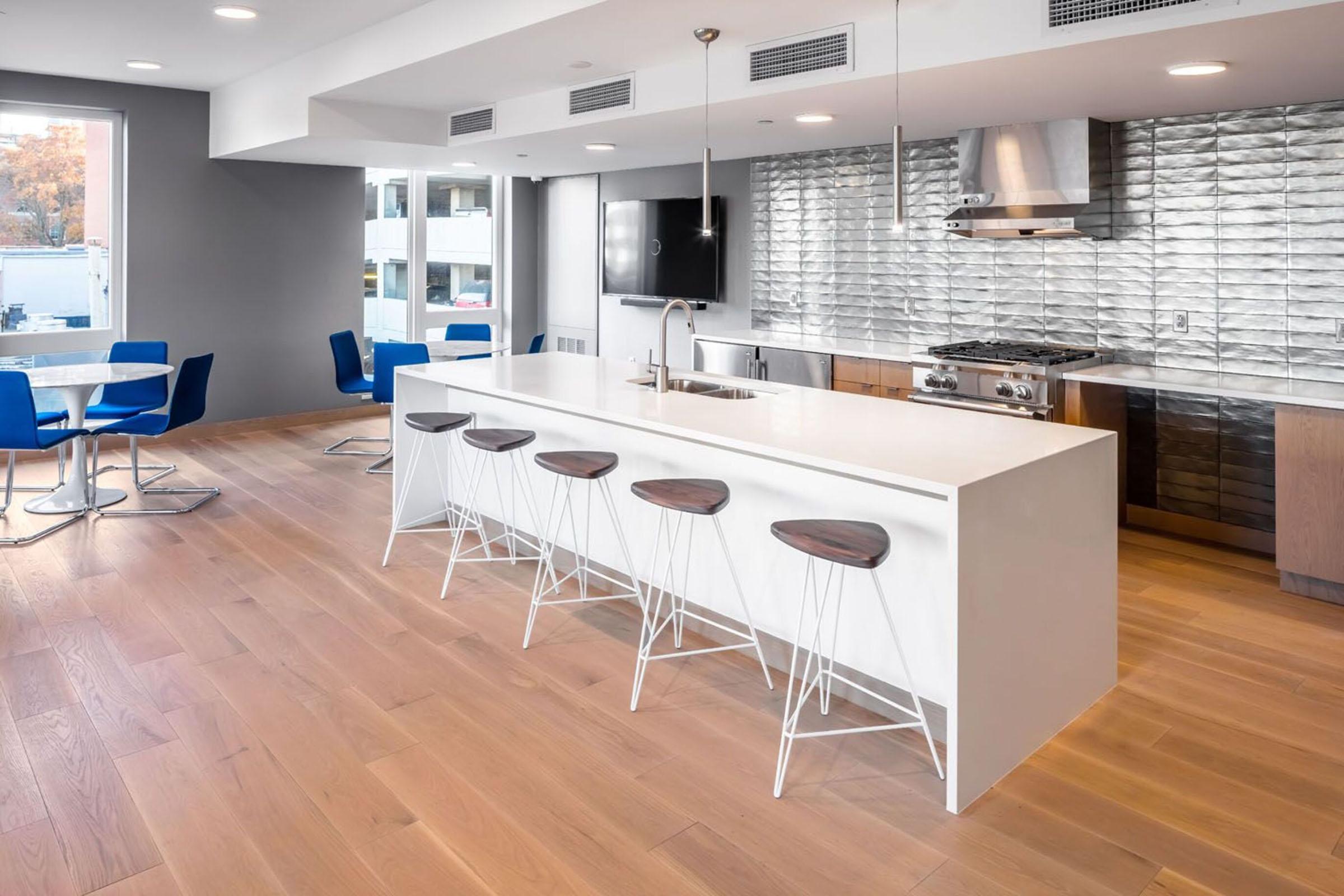 Modern kitchen interior featuring a long white island with five minimalist bar stools. The backdrop consists of a sleek, metallic wall. Natural light filters through large windows, illuminating blue chairs at a nearby dining table. The space has wooden flooring, creating a warm and inviting atmosphere.