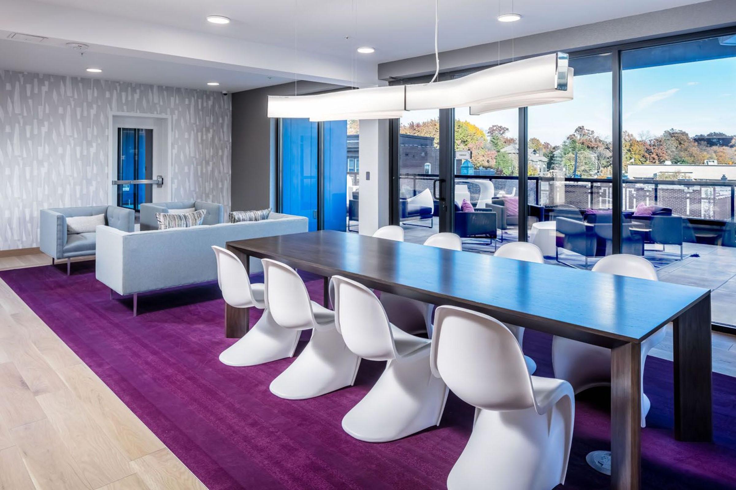 A modern dining area featuring a long wooden table surrounded by white chairs, situated on a purple carpet. Large windows allow natural light to fill the space, showcasing a view of outdoor scenery. Stylish sofas are positioned against the wall, enhancing the contemporary design.