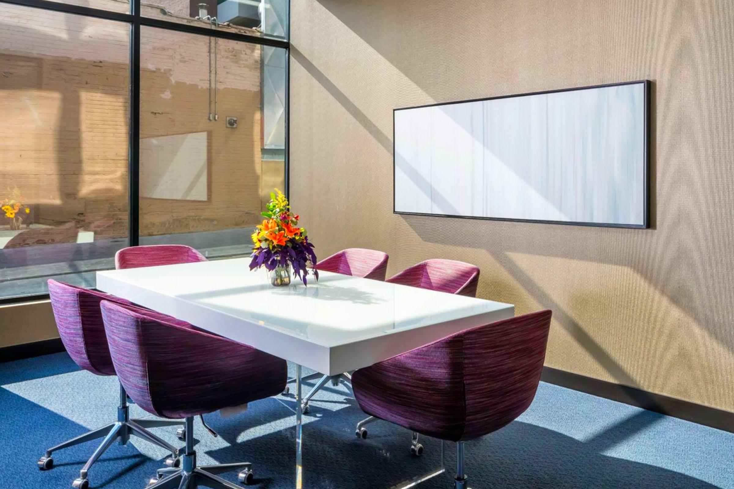 A modern conference room featuring a white table surrounded by purple ergonomic chairs. A vibrant flower arrangement sits at the center of the table. The room has large windows allowing natural light to fill the space, with a minimalistic wall art piece hanging on one side.