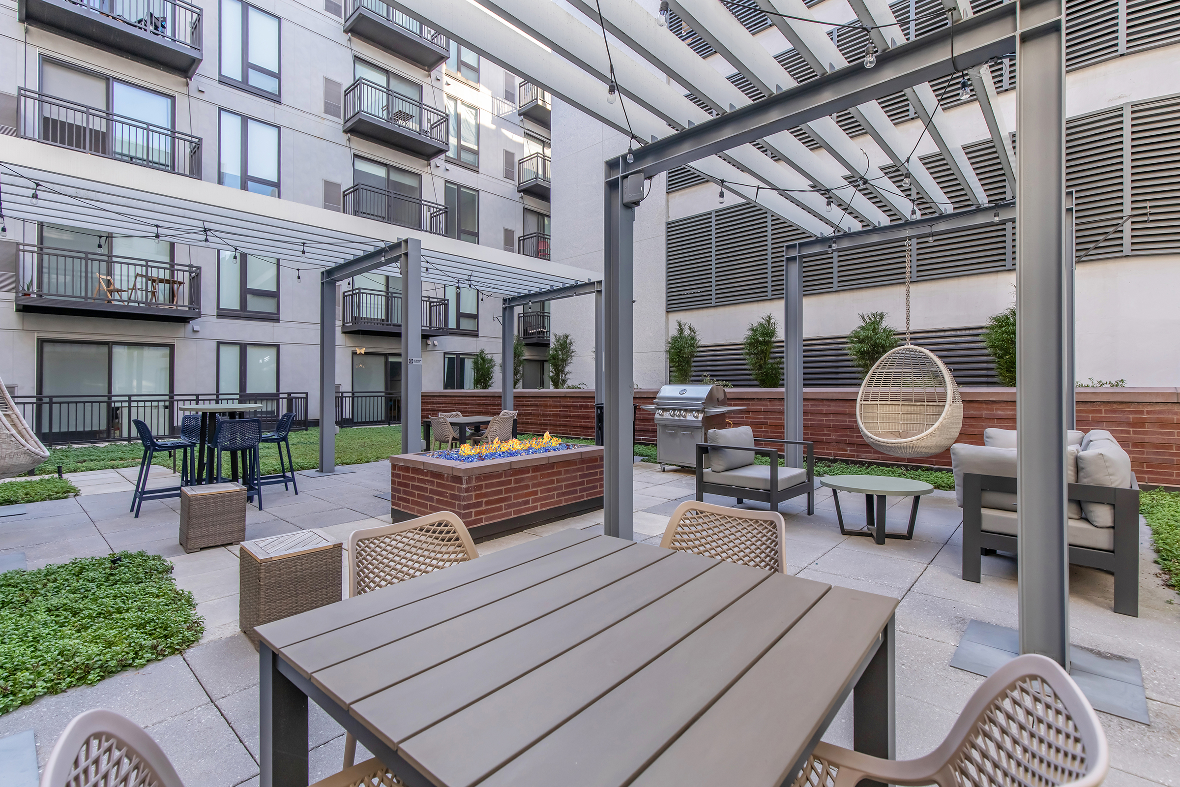 A modern outdoor courtyard featuring a dining table, hanging chairs, a grilling area, and a fire pit surrounded by greenery. The space is enclosed by apartment buildings with balconies, creating a cozy social environment.