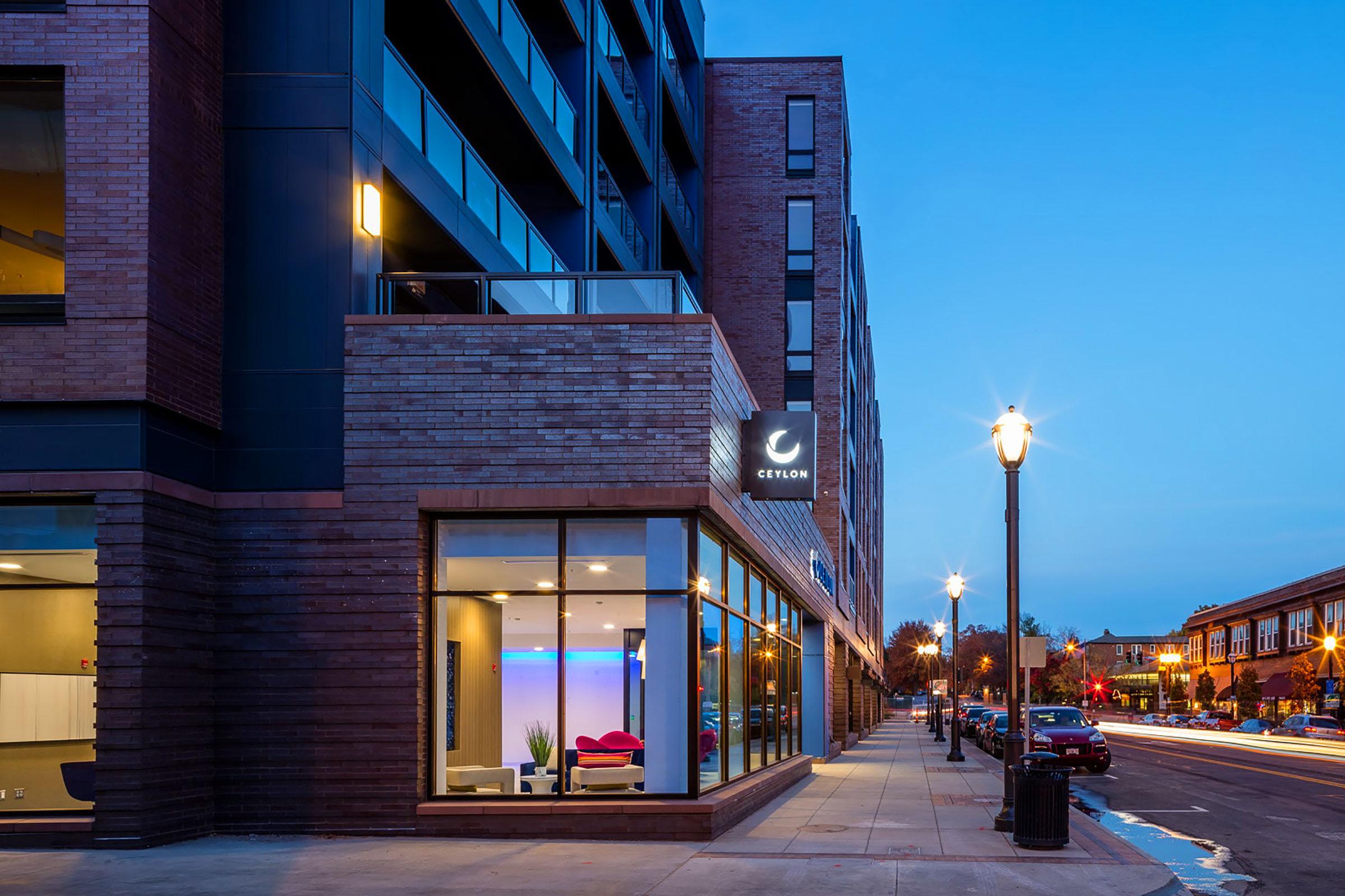 Modern building exterior at dusk, featuring large windows with interior lighting. A sleek sign with the name "Ceylon" is visible. The sidewalk is lined with street lamps, and the street is partly empty, showcasing a tranquil urban environment.