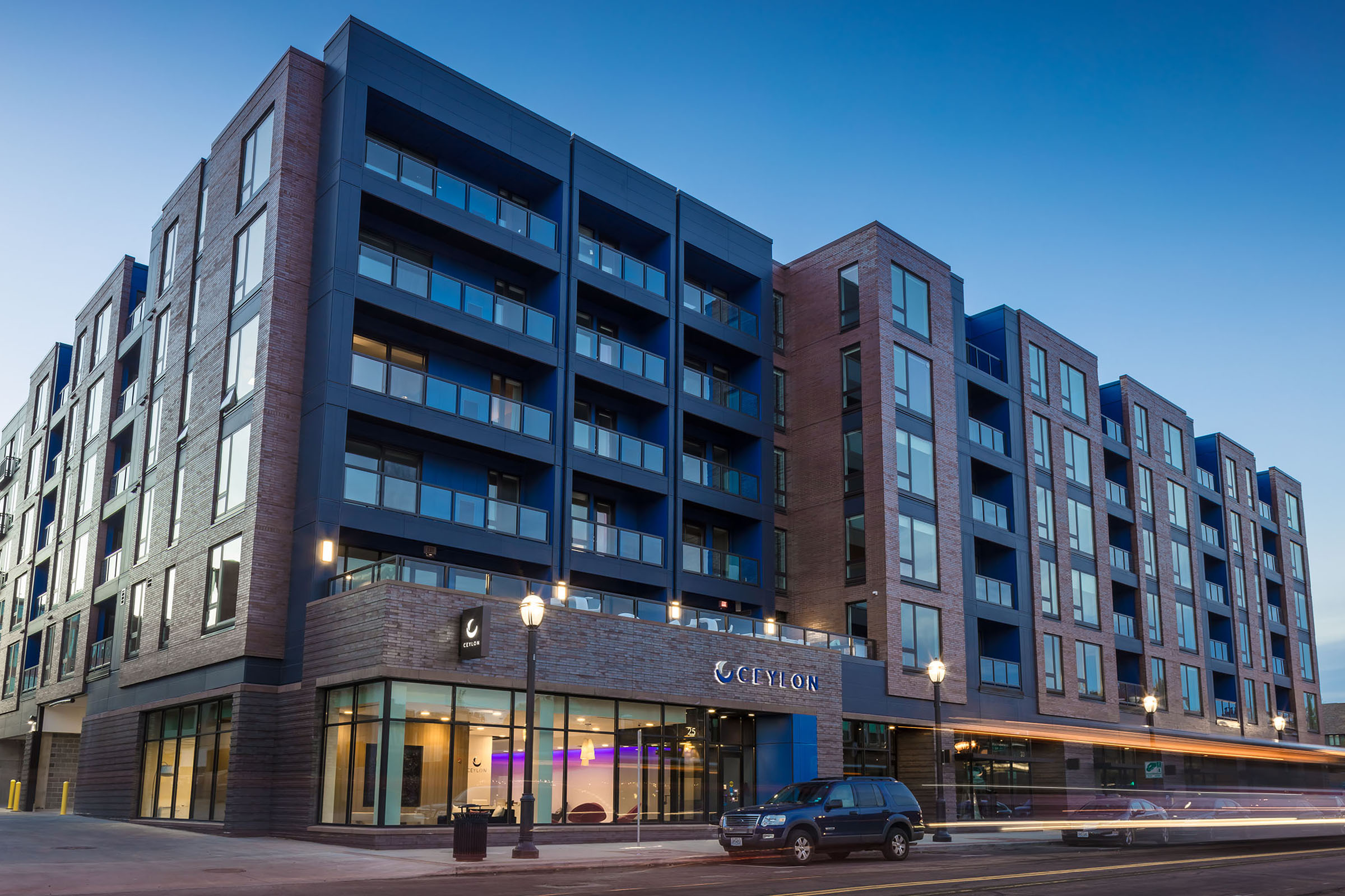 Modern apartment building with multiple stories. The façade features a mix of dark and light bricks, large windows, and a prominent entrance labeled "CEYLON." Streetlights illuminate the exterior, and a parked car is visible in front, with a twilight sky creating a serene atmosphere.
