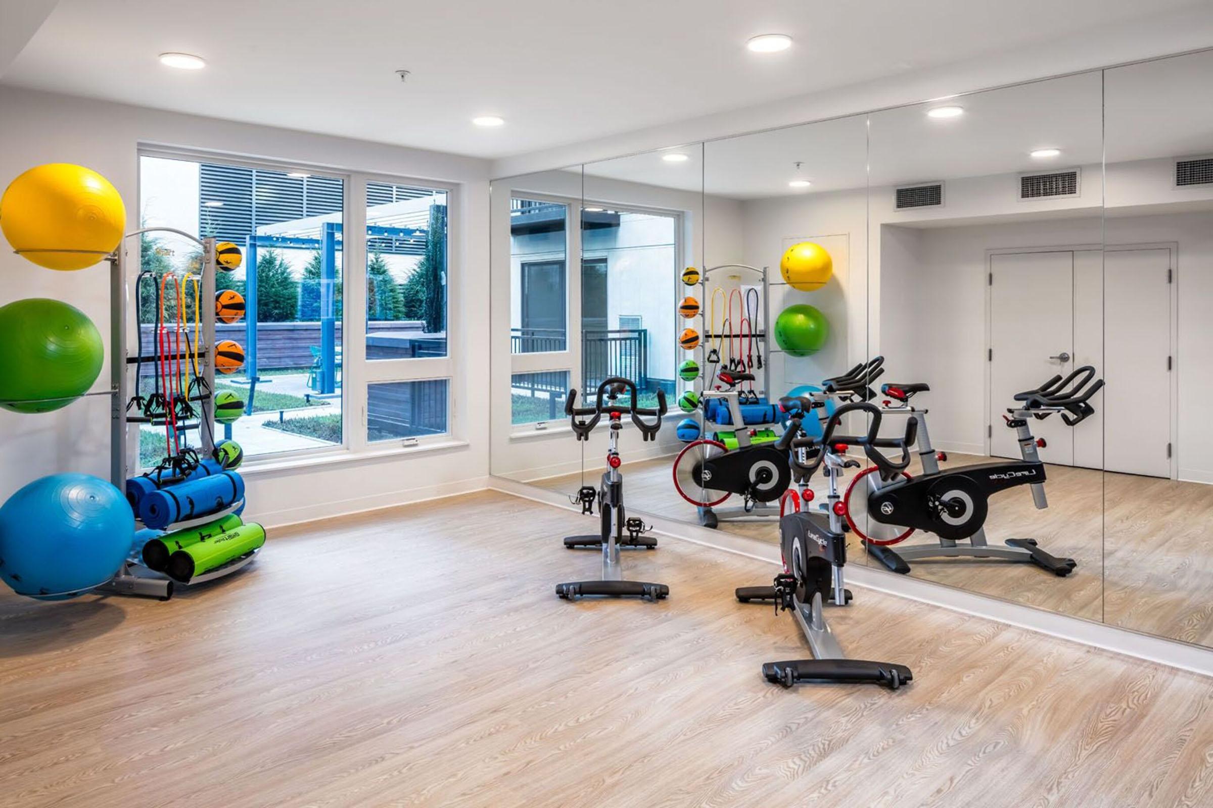 A modern fitness room featuring exercise bikes, mats, and various exercise balls. Mirrors on one wall reflect the bright, well-lit space, which includes equipment neatly organized for workouts. Large windows let in natural light, enhancing the overall ambiance.