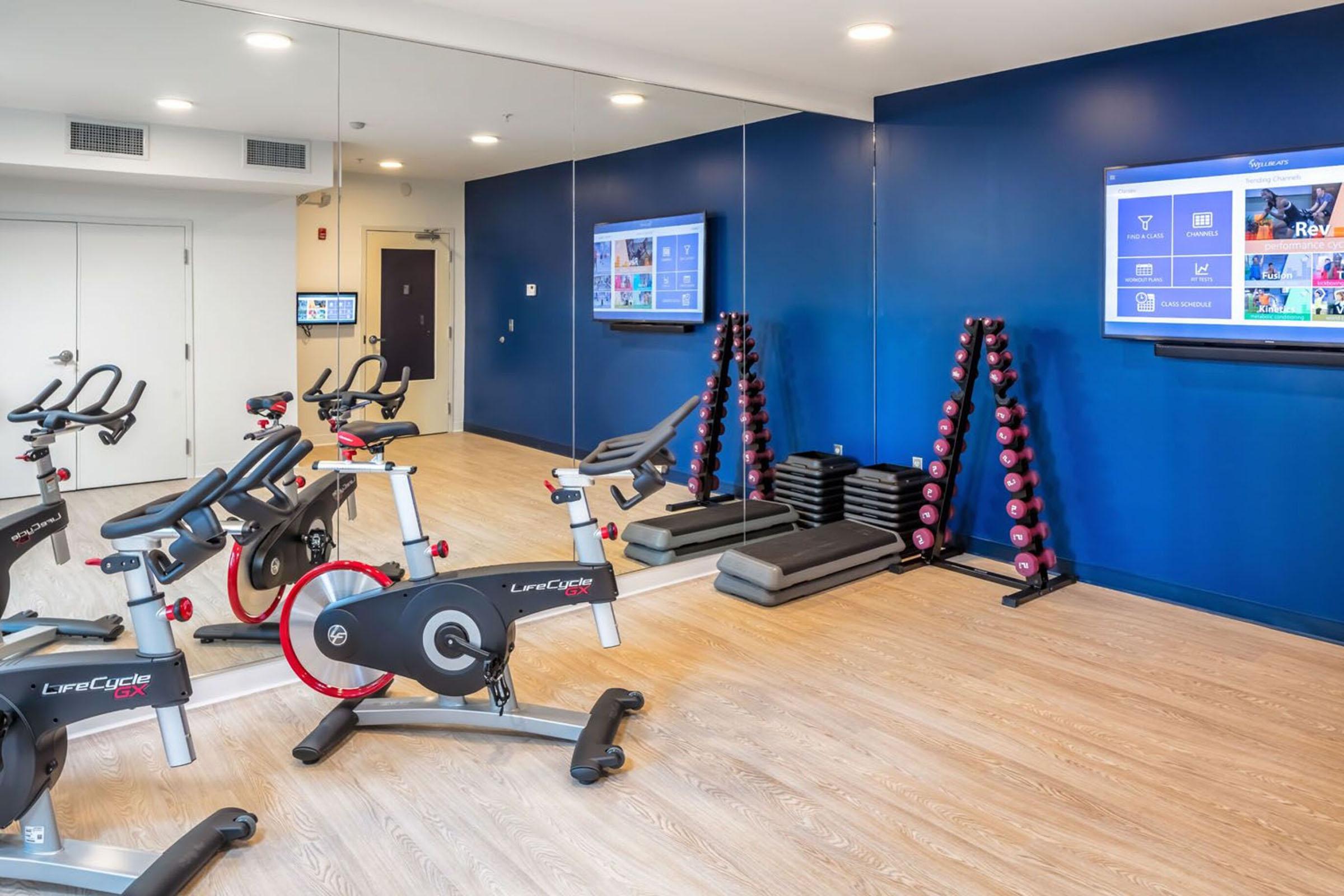 A modern fitness room featuring several stationary bikes, a set of dumbbells arranged on a rack, exercise mats, and two large screens displaying fitness information. The walls are painted blue, and there are mirrors enhancing the spacious feel of the room.