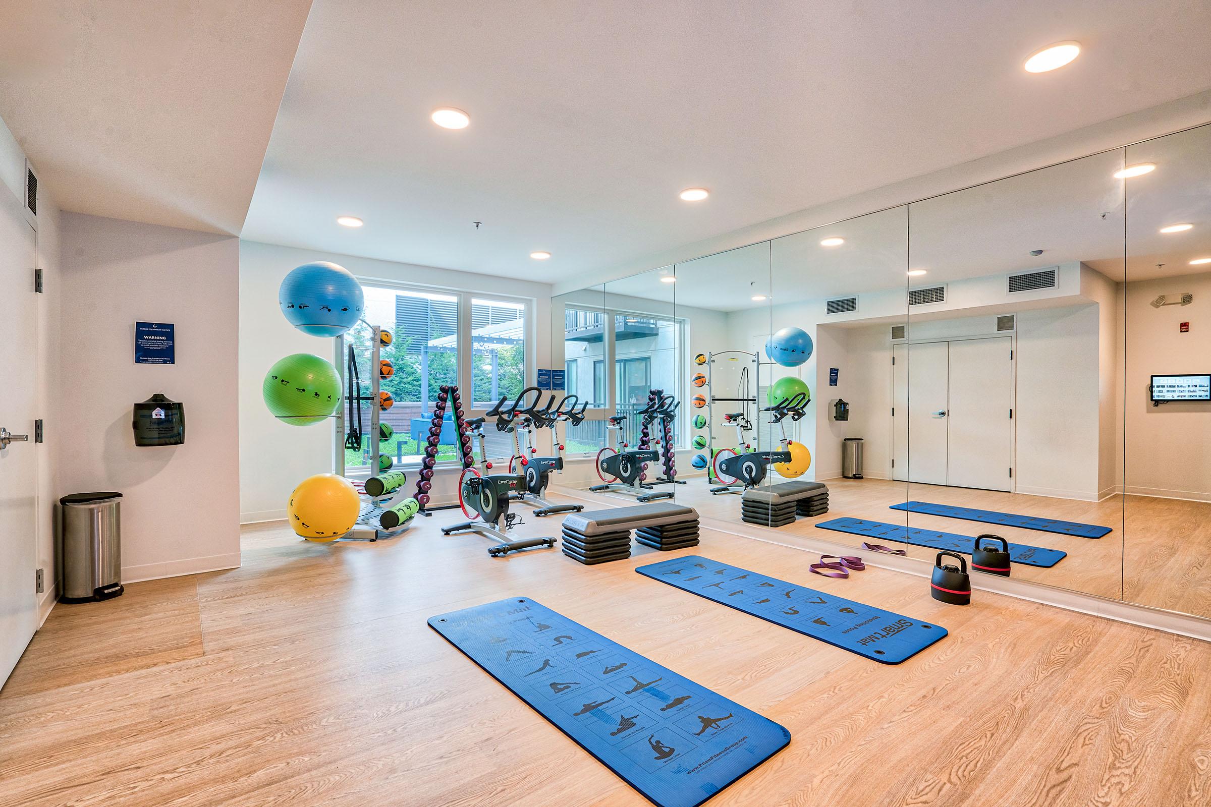 A modern gym interior featuring exercise mats, free weights, exercise bikes, and colorful exercise balls. Large mirrors line one wall, enhancing the sense of space. The room is well-lit and designed for various fitness activities, creating an inviting atmosphere for workouts.