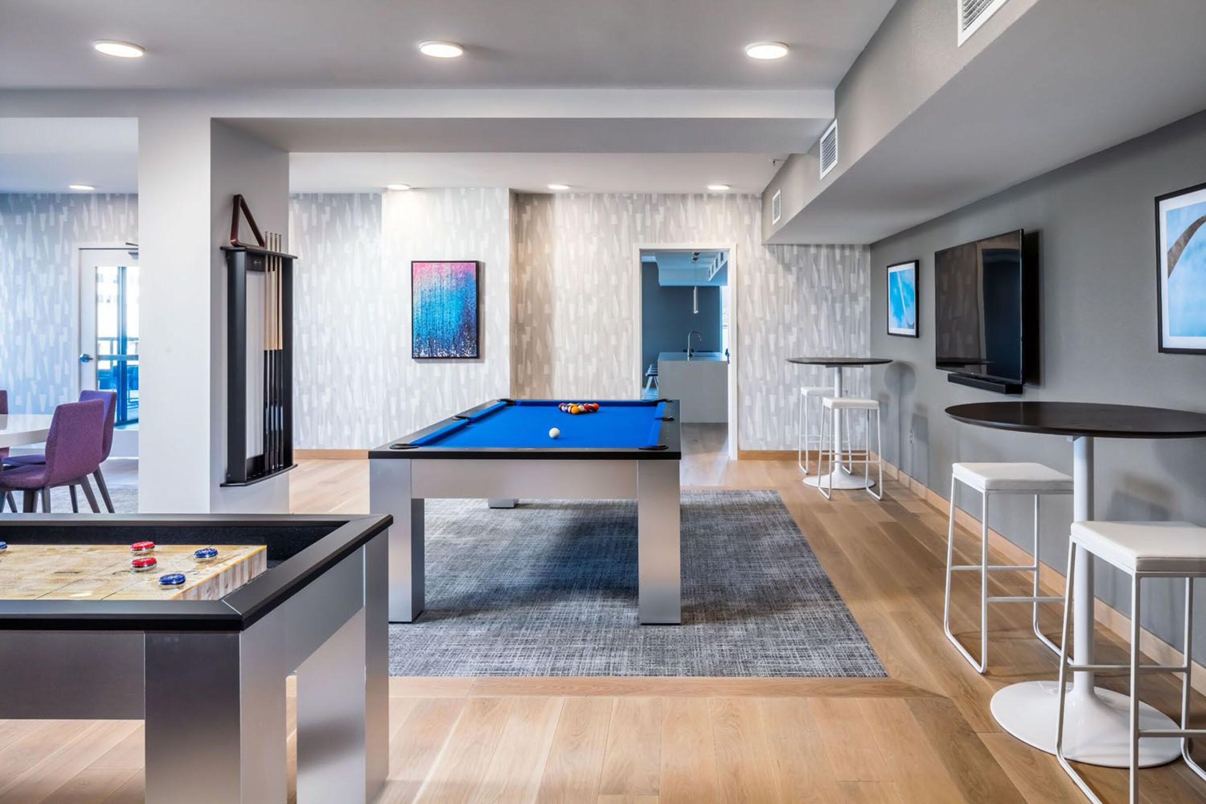 Interior of a modern game room featuring a blue pool table, a tabletop game with colorful pieces, and seating areas. There are flat-screen TVs mounted on the walls, stylish light fixtures, and contemporary decor with a light color scheme, creating a vibrant and inviting atmosphere for relaxation and socializing.