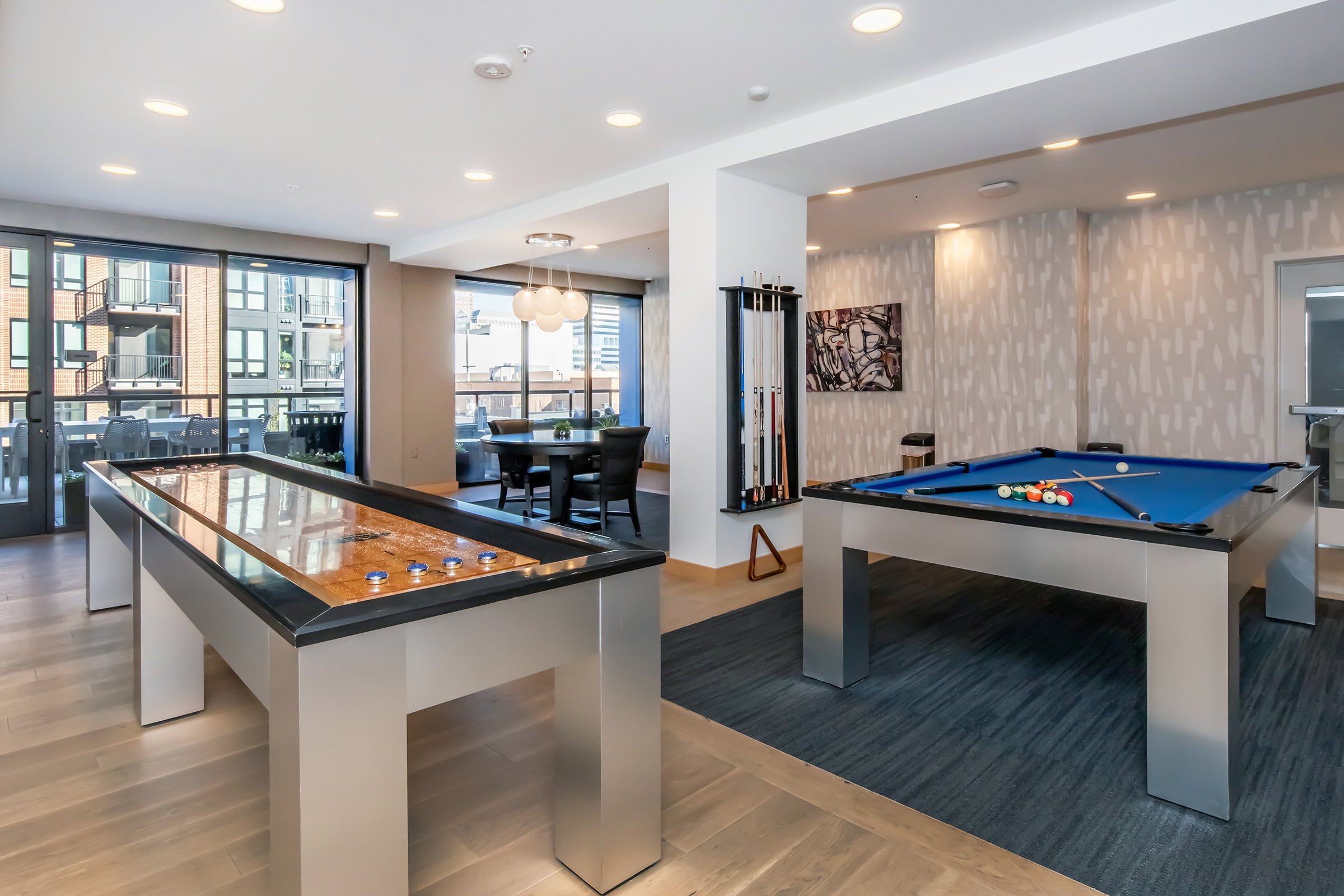 Interior of a modern recreational room featuring a billiards table and a shuffleboard table. Natural light streams in through large windows, illuminating the space decorated with contemporary artwork. Stylish seating is visible in the background, creating a welcoming atmosphere for leisure activities.