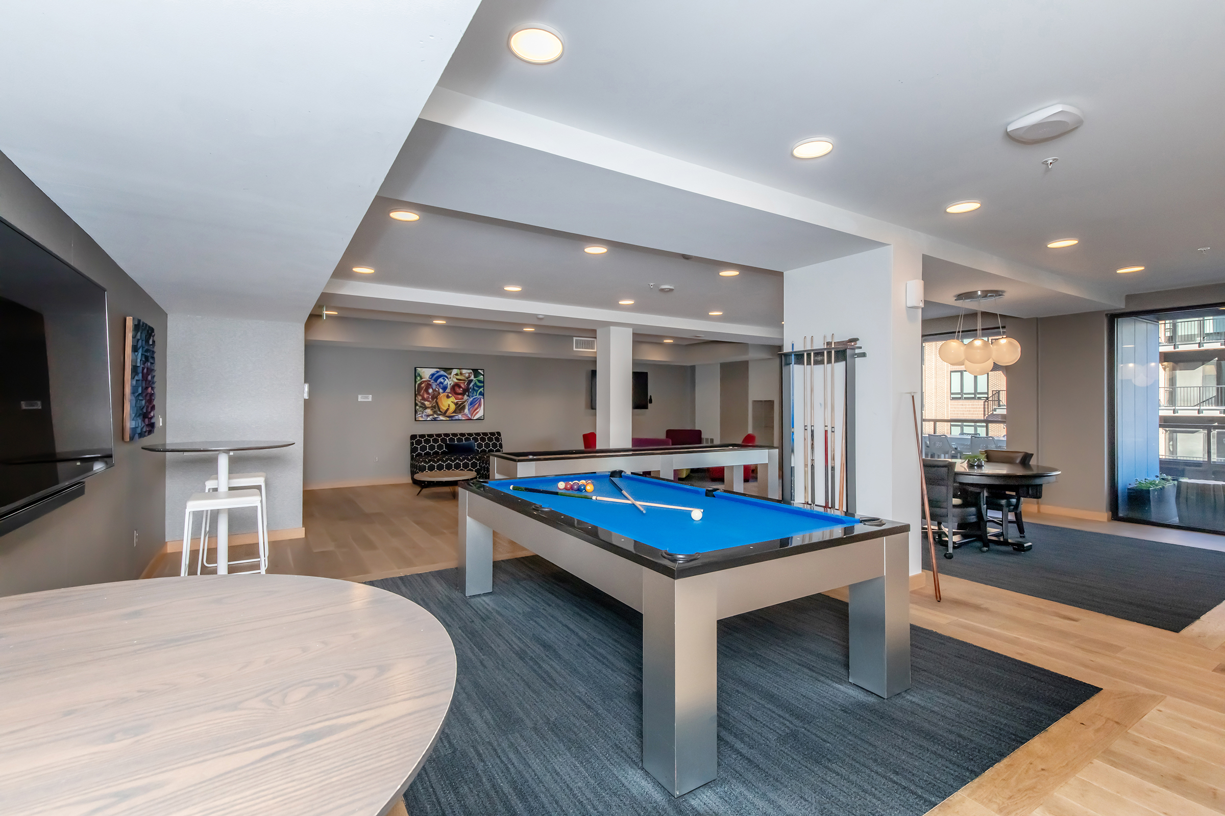 A spacious game room featuring a blue pool table, wooden flooring, and modern decor. There are comfortable seating areas, wall art, and a flat-screen TV visible. Bright overhead lights illuminate the space, creating a welcoming atmosphere for leisure activities.
