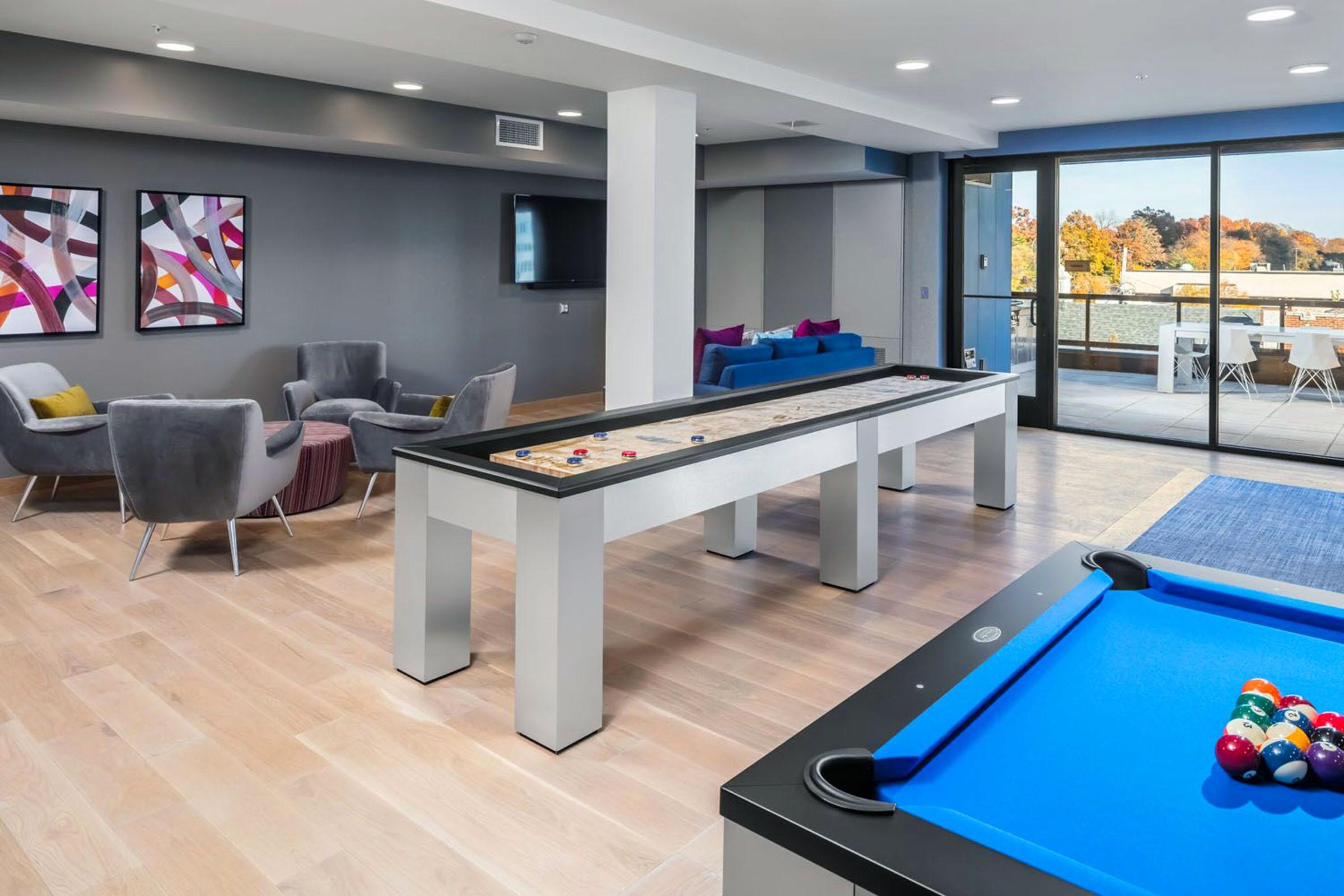 Modern indoor space featuring a stylish lounge area with gray chairs and colorful cushions, a shuffleboard table in the center, and a blue pool table in the foreground. Large windows offer a view of the outdoors, with access to a balcony. Abstract artwork adorns the walls, adding a contemporary touch to the decor.