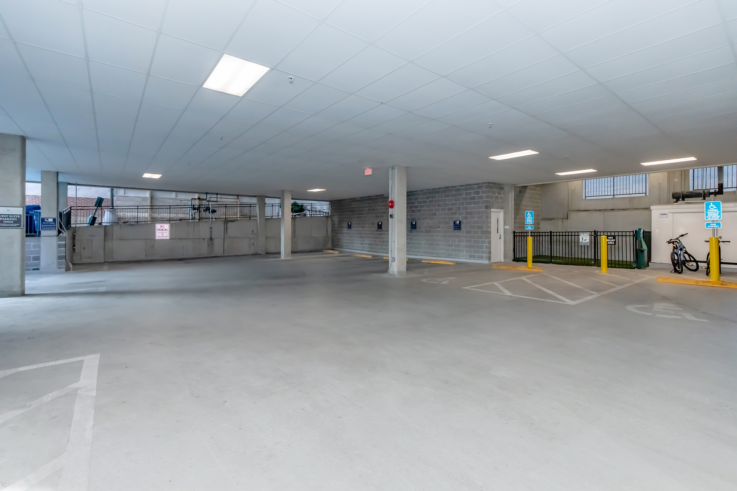 An empty indoor parking garage with concrete floors, illuminated by ceiling lights. The space features yellow parking barriers, bike racks, and security signage. A garage door is visible at the far end, with a few windows allowing natural light to enter.
