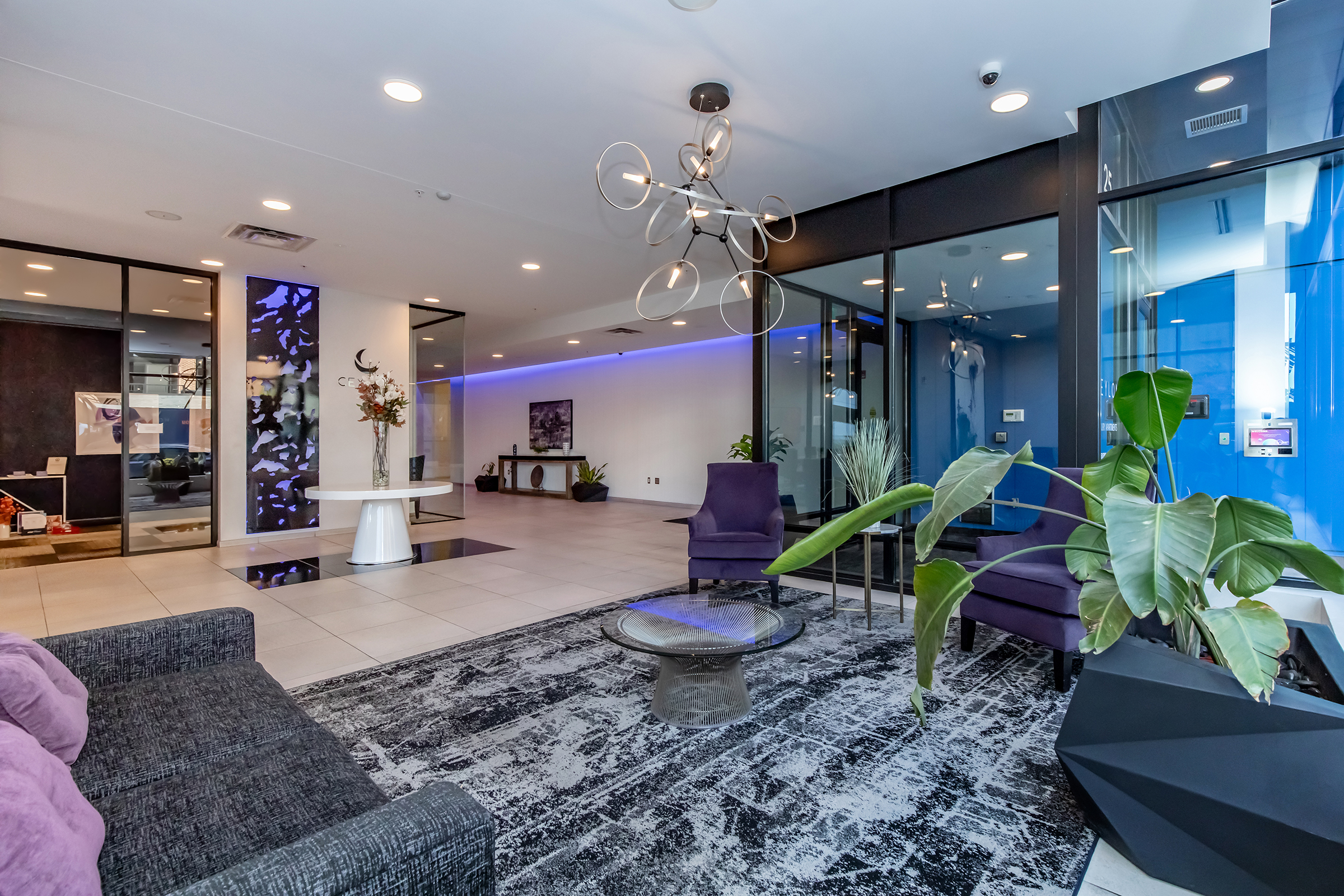 Modern lobby area featuring sleek furniture, including a dark sofa and purple chairs, a decorative plant, and a contemporary light fixture. Glass walls create an open atmosphere, enhanced by blue ambient lighting. A stylish rug completes the aesthetic, emphasizing a chic and inviting design.