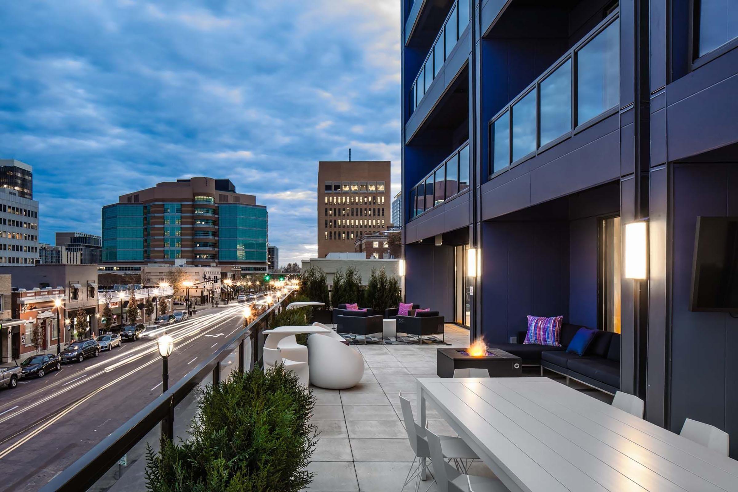 A modern rooftop terrace featuring a sleek table and seating area, surrounded by potted plants. In the background, city buildings are visible under a cloudy sky, with street lights illuminating a busy road. The scene captures an inviting atmosphere for outdoor gatherings or relaxation.