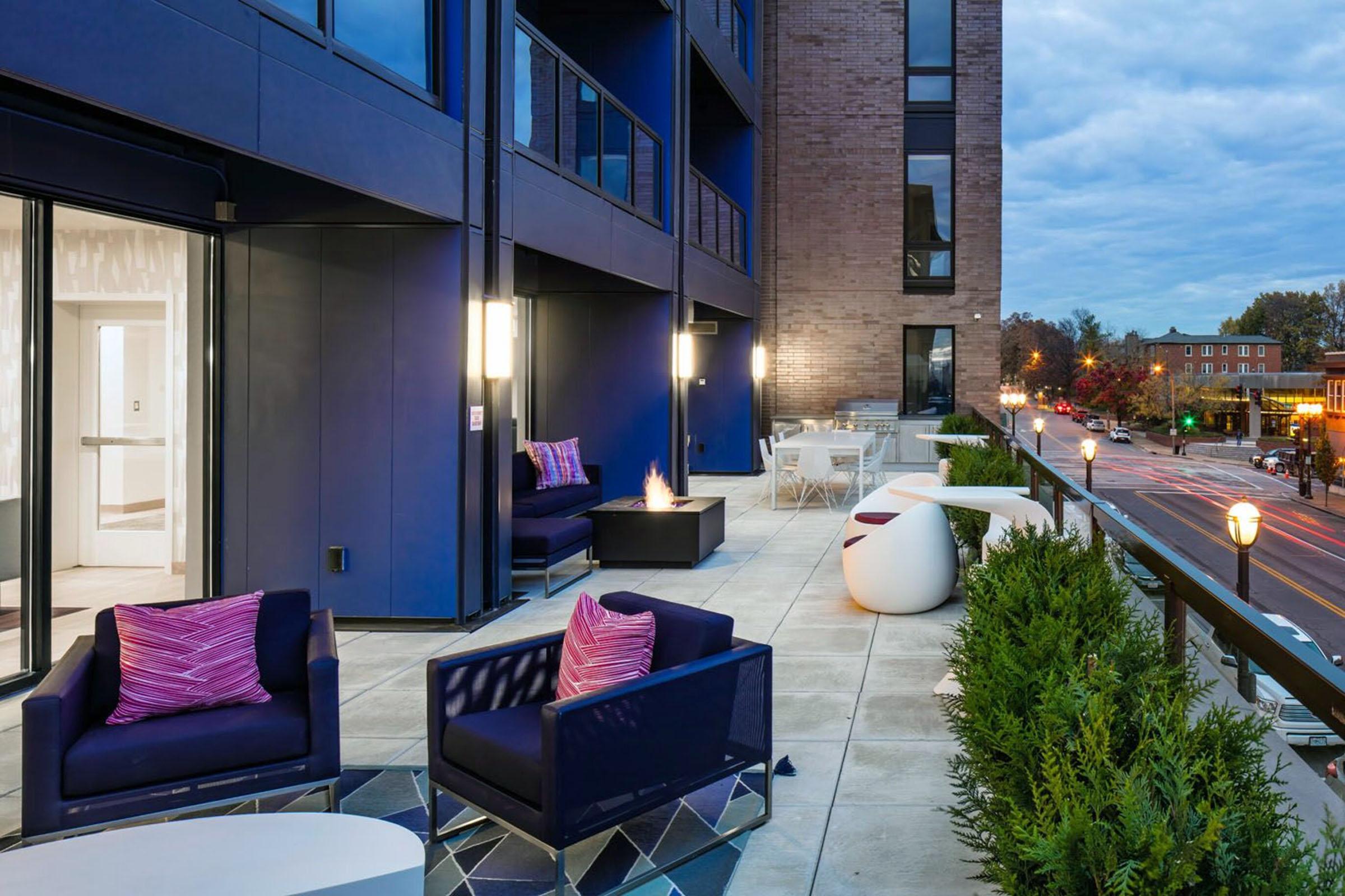 A modern outdoor terrace with stylish furniture, including purple and white chairs, a small fire pit, and a glass railing. The backdrop features a contemporary building with large windows and a sidewalk lined with trees and streetlights, under a cloudy sky.