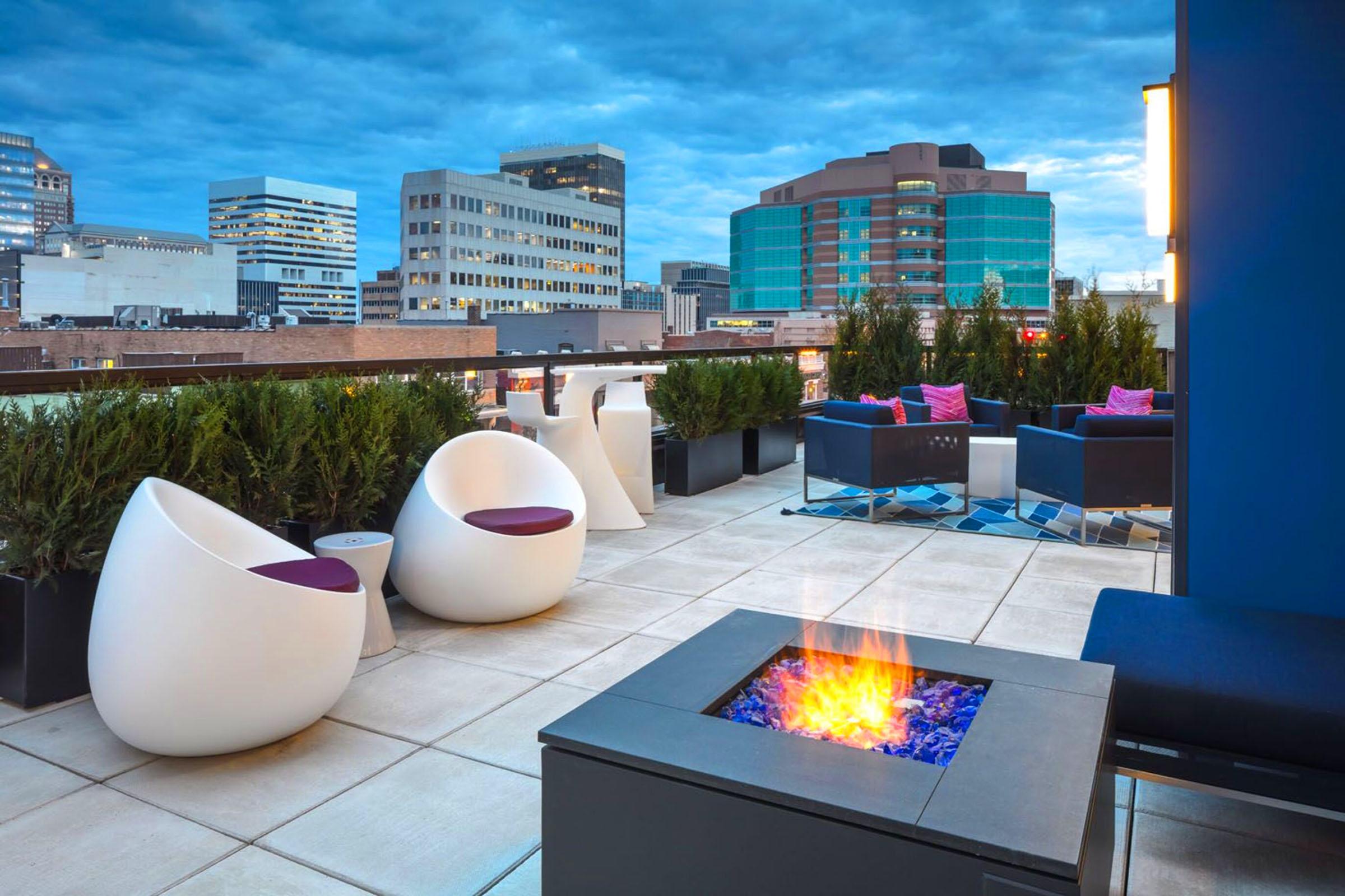 A modern rooftop patio with stylish white chairs and black seating. A fire pit with blue stones creates a cozy atmosphere. Lush green plants line the area, and the backdrop showcases a city skyline under a cloudy blue sky.