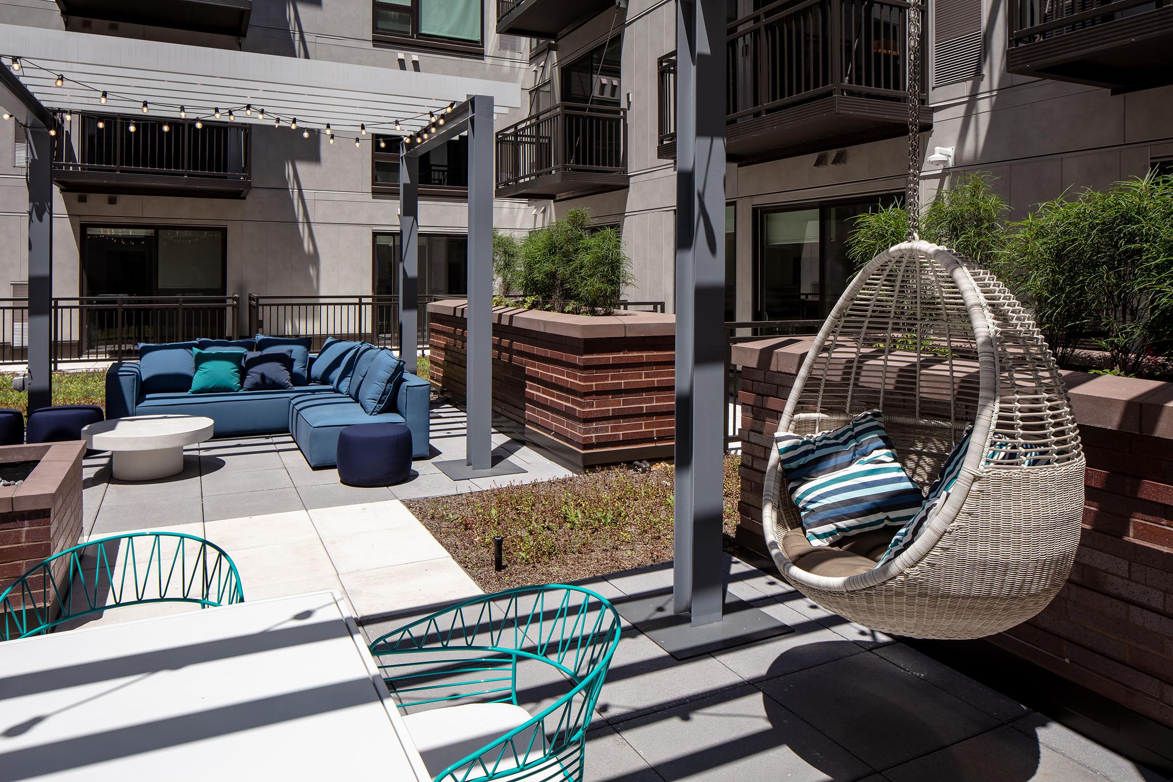 A modern outdoor lounge area featuring a cozy blue sectional sofa, a hanging chair, and a dining table. The space is decorated with plants and string lights, offering a relaxed atmosphere in an apartment complex courtyard.