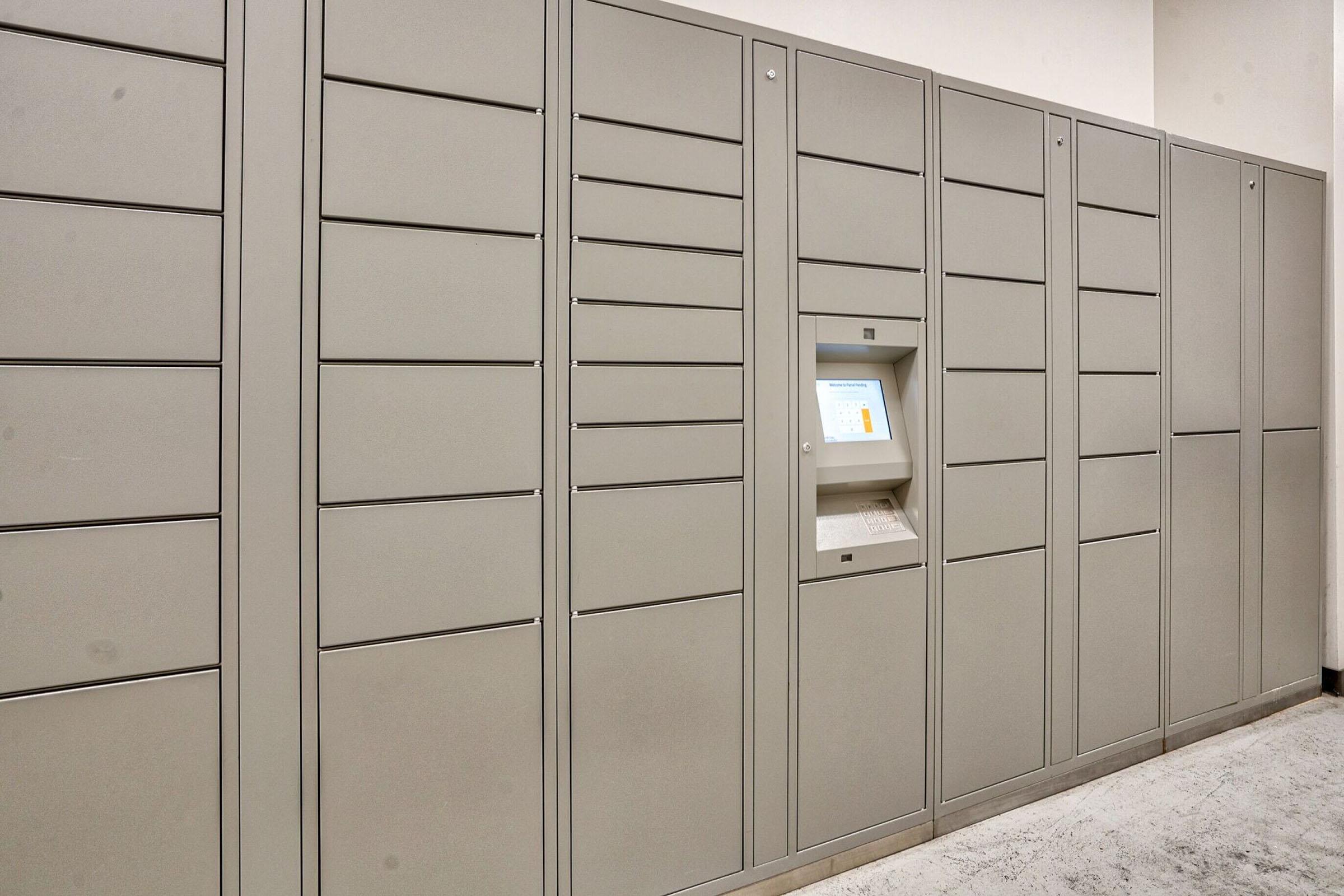 A row of gray, metallic lockers with a digital touchscreen interface integrated into the center. The lockers are empty, featuring horizontal slits for access, set against a neutral-colored wall. The floor appears to be concrete, creating a minimalistic and utilitarian design.