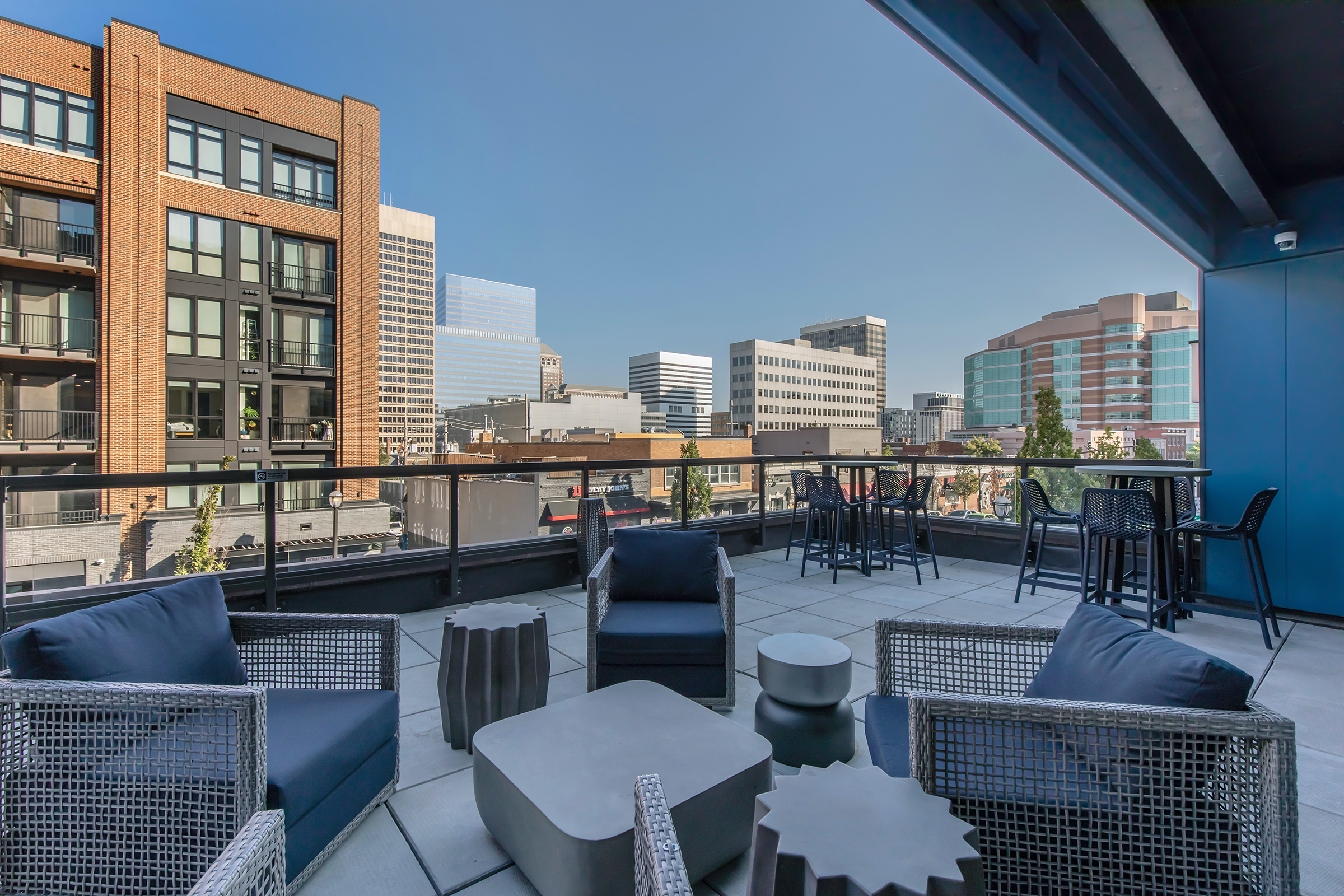 A modern rooftop terrace featuring stylish outdoor furniture, including armchairs and a table, overlooking a vibrant city skyline with a mix of high-rise buildings and clear blue skies. Perfect for social gatherings or relaxation.