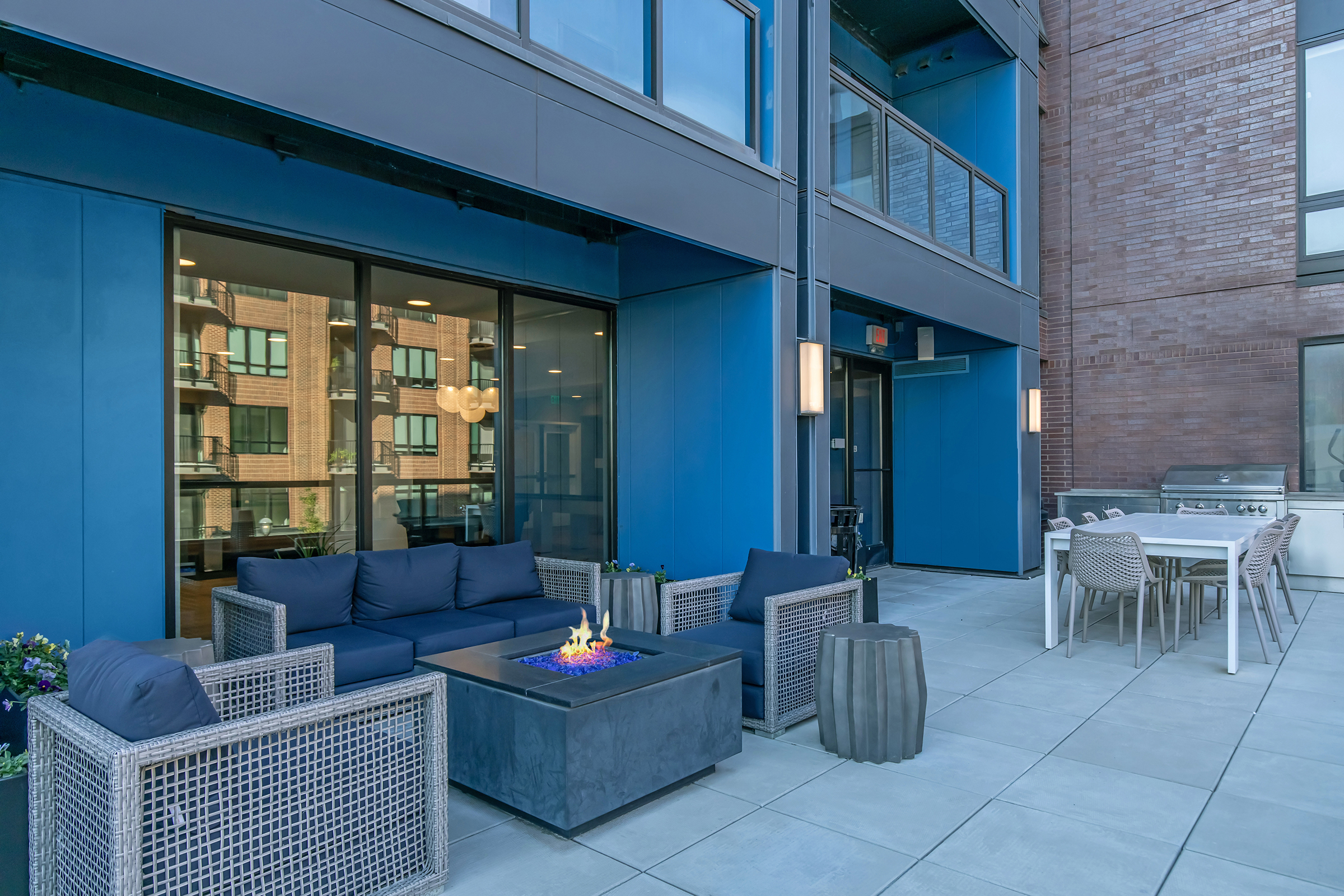 A modern outdoor patio featuring a seating area with navy blue cushions, a fire pit table with a flame, and a dining table with chairs. The backdrop includes large windows showcasing an urban view and contemporary architecture in a vibrant color scheme.