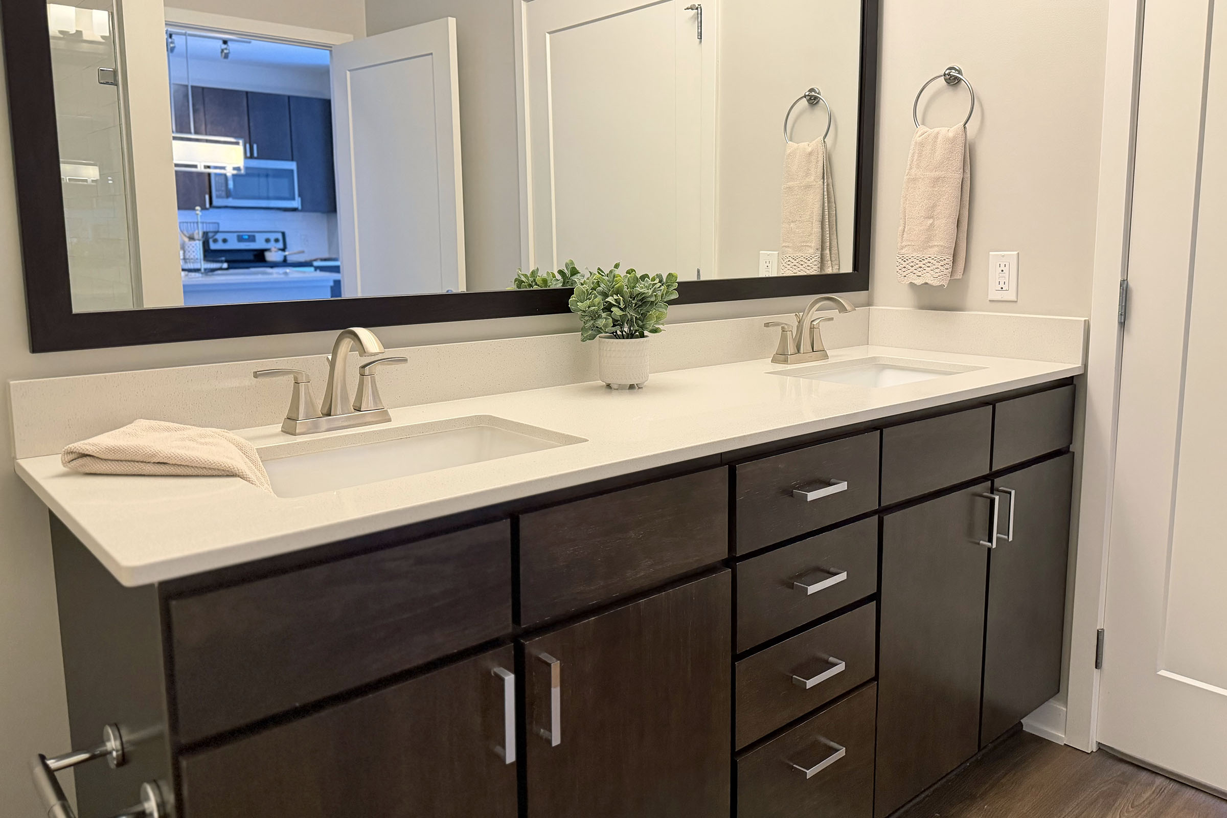 A modern bathroom with a double vanity sink. The countertop is light-colored with two undermount sinks and stylish faucets. Dark wood cabinets provide storage underneath, and there are two folded towels hanging on rings above. A small potted plant adds a decorative touch, and a doorway leads to another room in the background.