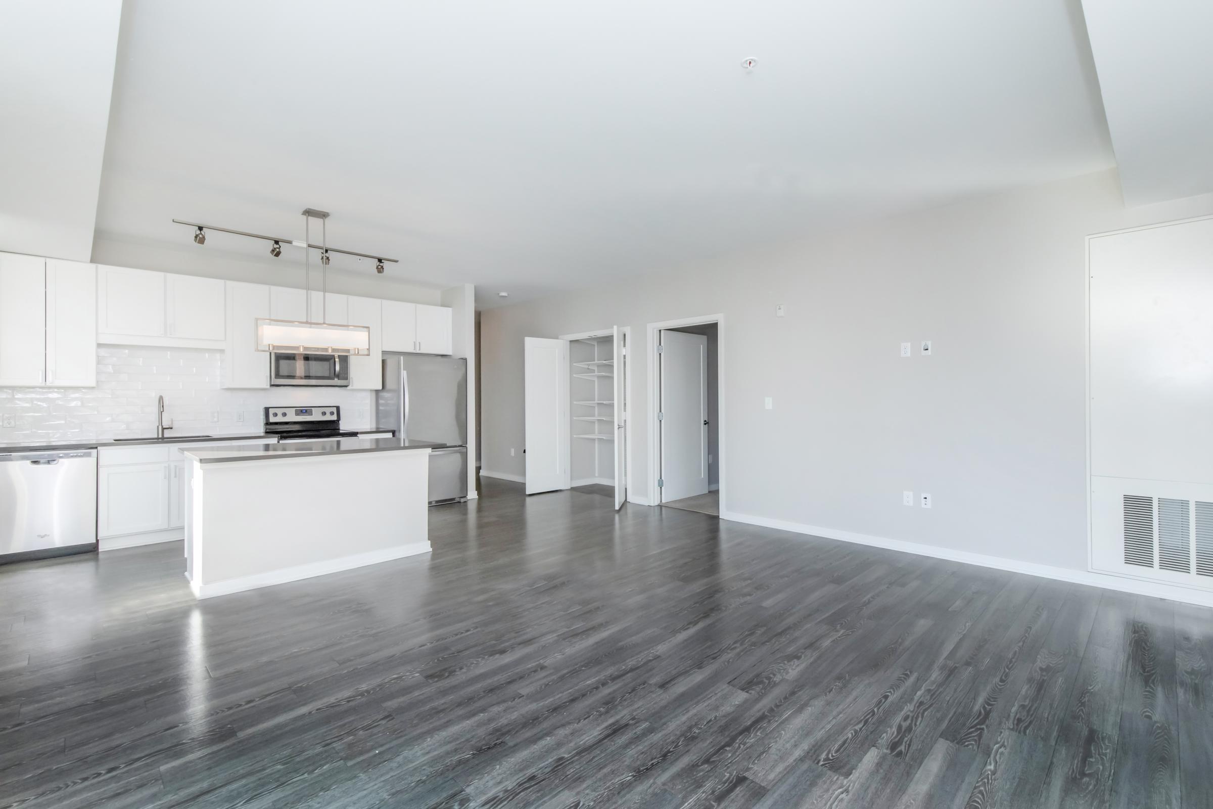 Modern open-concept kitchen and living area featuring sleek white cabinetry, stainless steel appliances, and dark hardwood floors. The space is well-lit with large windows and includes a door leading to another room, showcasing a minimalist and contemporary design.