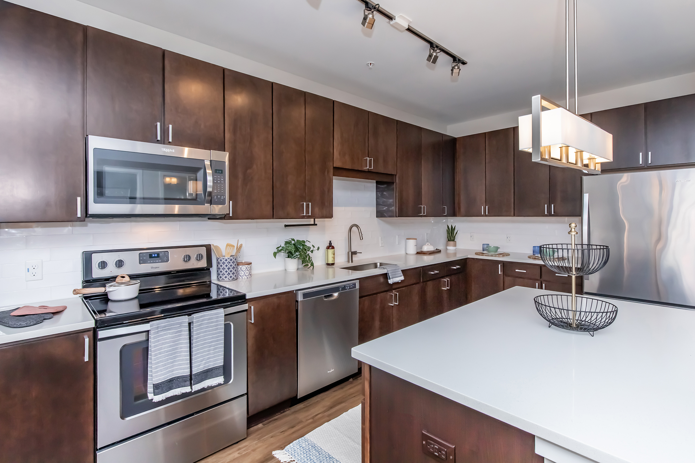 A modern kitchen featuring dark wooden cabinets, stainless steel appliances, a large island with a white countertop, and sleek lighting. The space is well-organized with a microwave, stove, sink, and decorative plants, creating a warm and inviting atmosphere.