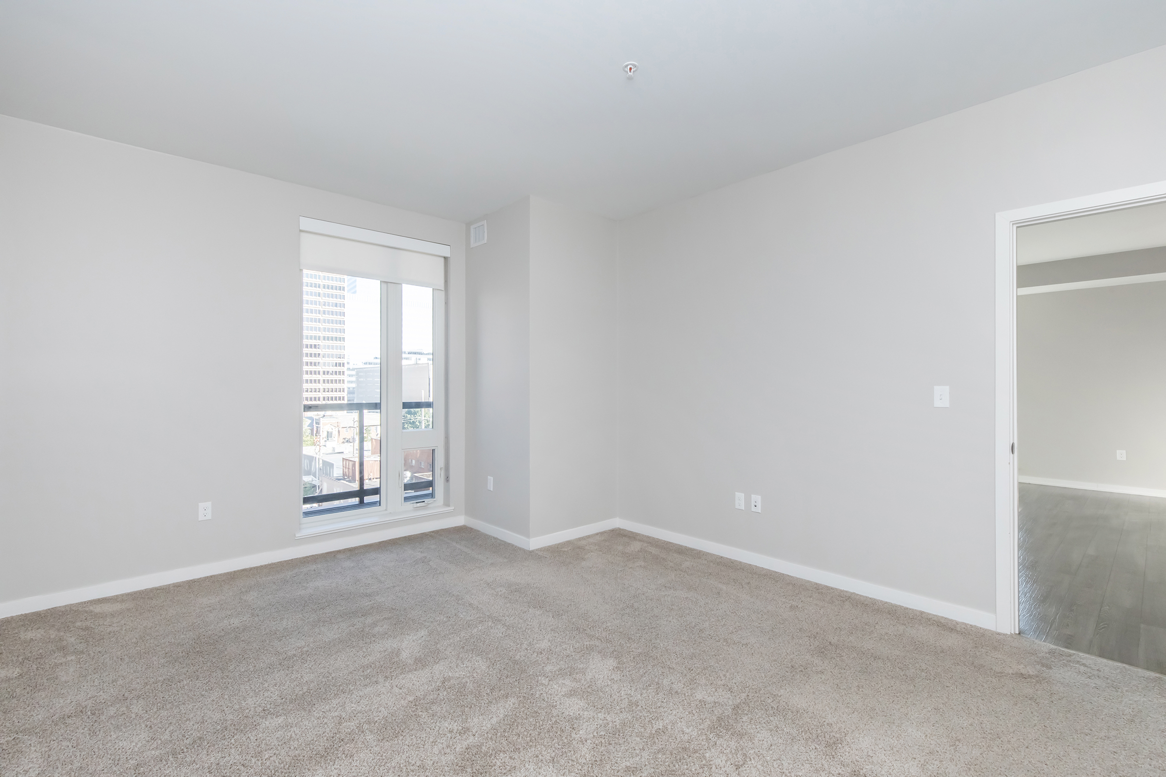 Empty room with light gray walls and beige carpet. A large window lets in natural light, offering a view of a cityscape. There is a door on the right leading to another space, creating an open and spacious feel. The overall atmosphere is bright and modern.