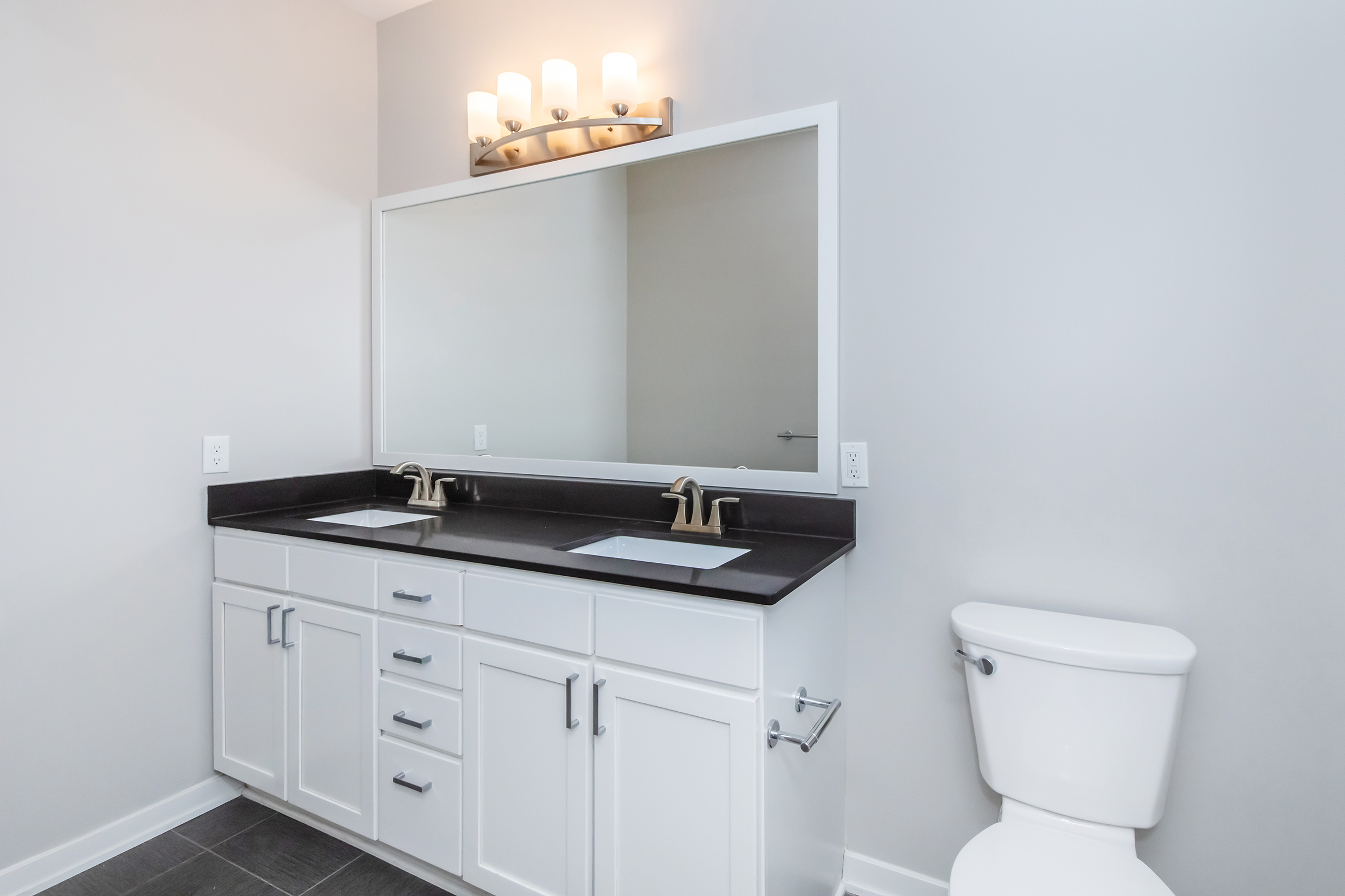 Modern bathroom featuring a double sink vanity with a dark countertop, large wall mirror, and contemporary light fixtures above. There is a white toilet to the right, and light gray walls complement the overall design, creating a clean and spacious aesthetic.