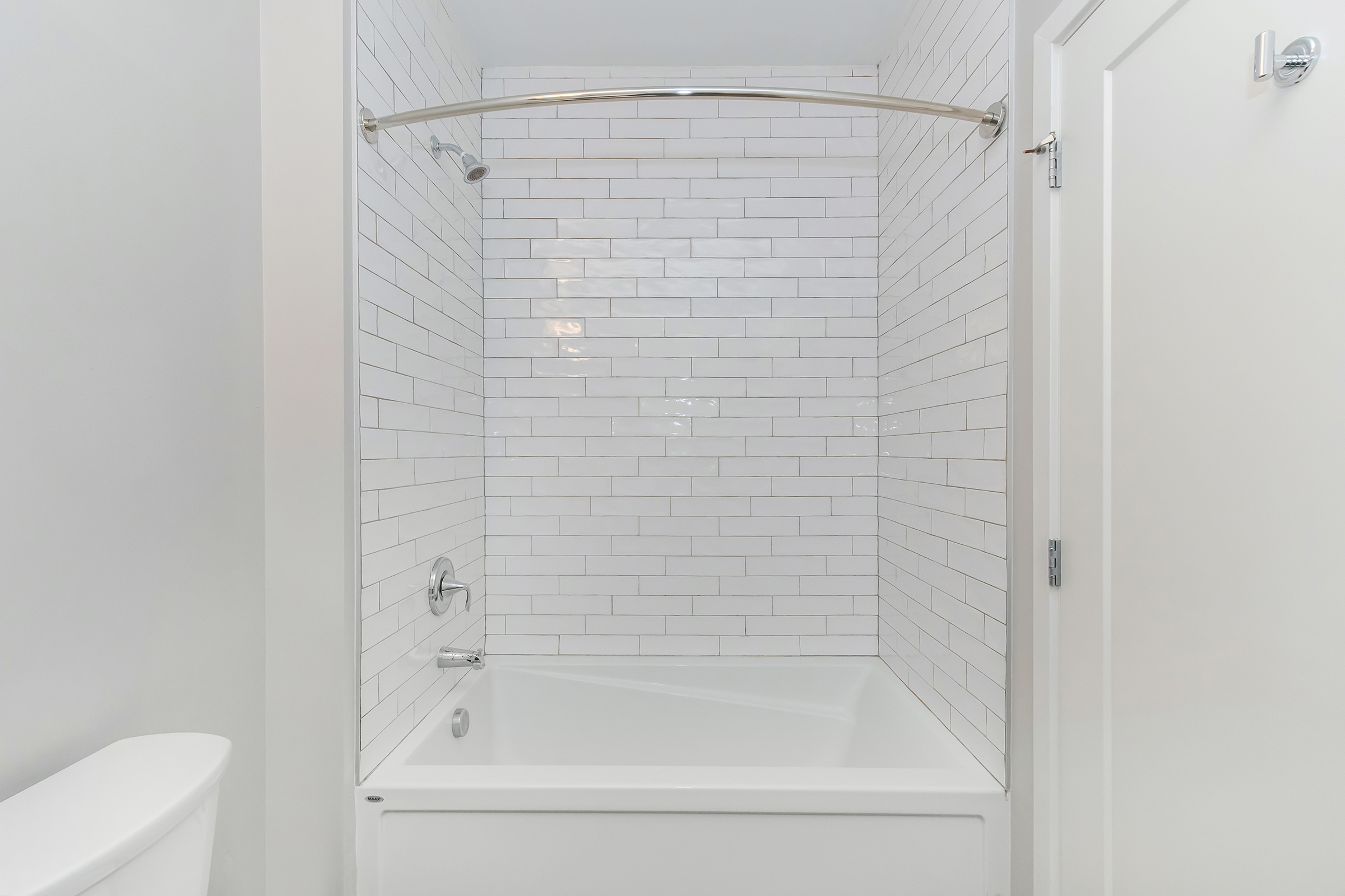 A clean and modern bathroom featuring a white tiled shower enclosure with a bathtub. The walls are covered in glossy white subway tiles, and there is a chrome shower rod. A simple white toilet is positioned to the left, contributing to the overall bright and minimalistic design.